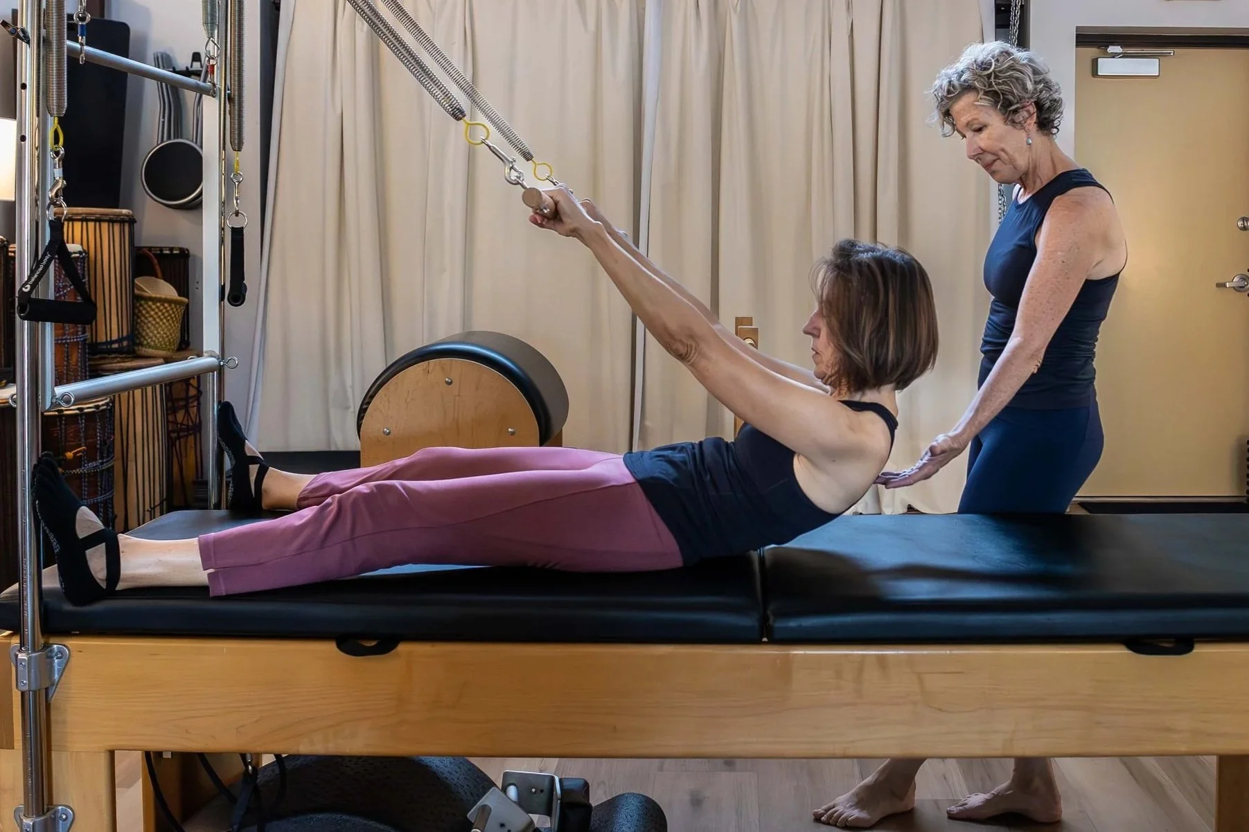 A woman in a black tank top and pink pants performs Pilates on a reformer machine with assistance from an instructor in a therapy or Pilates studio.