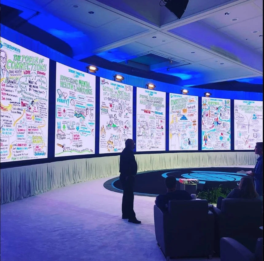 A large conference room with a curved stage and multiple digital screens displaying colorful handwritten notes and illustrations. Several people are seated and standing, observing the presentation in a dimly lit room with blue lighting.