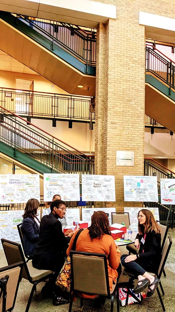 A group of people seated around a table engaged in discussion. Behind them are poster boards with various notes and illustrations. The setting appears to be an indoor space with staircases and a brick column.