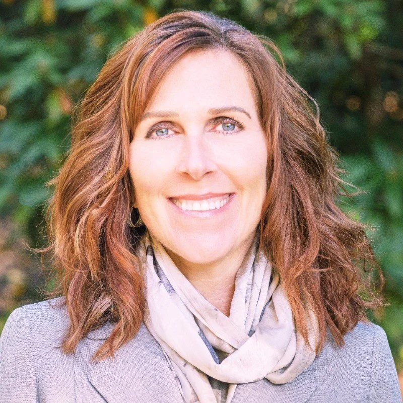 A woman with wavy, shoulder-length brown hair and blue eyes, smiling and wearing a light gray blazer and a light-colored scarf with dark accents, standing outdoors with greenery in the background.