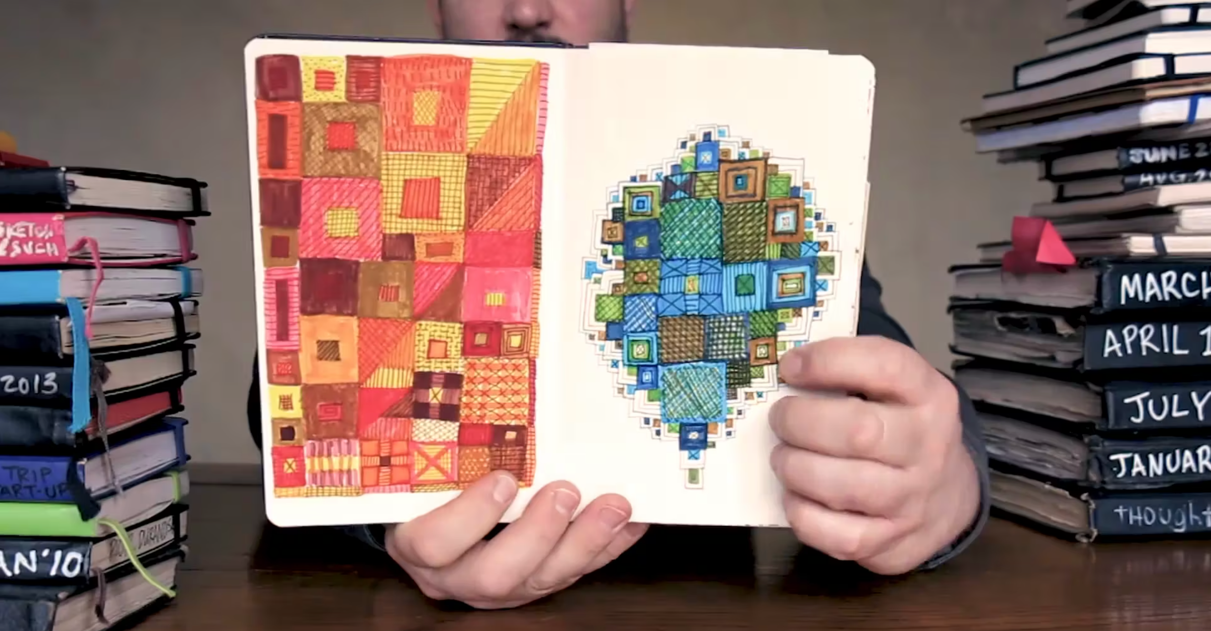 Person holding a sketchbook open to two pages featuring colorful geometric patterns in red, orange, blue, and green. Stacks of sketchbooks are labeled with months and years in the background.