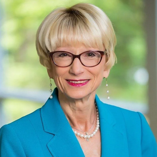 A woman with short blonde hair, wearing glasses, a turquoise blazer, pearl earrings and necklace, smiling in front of a blurred green background.