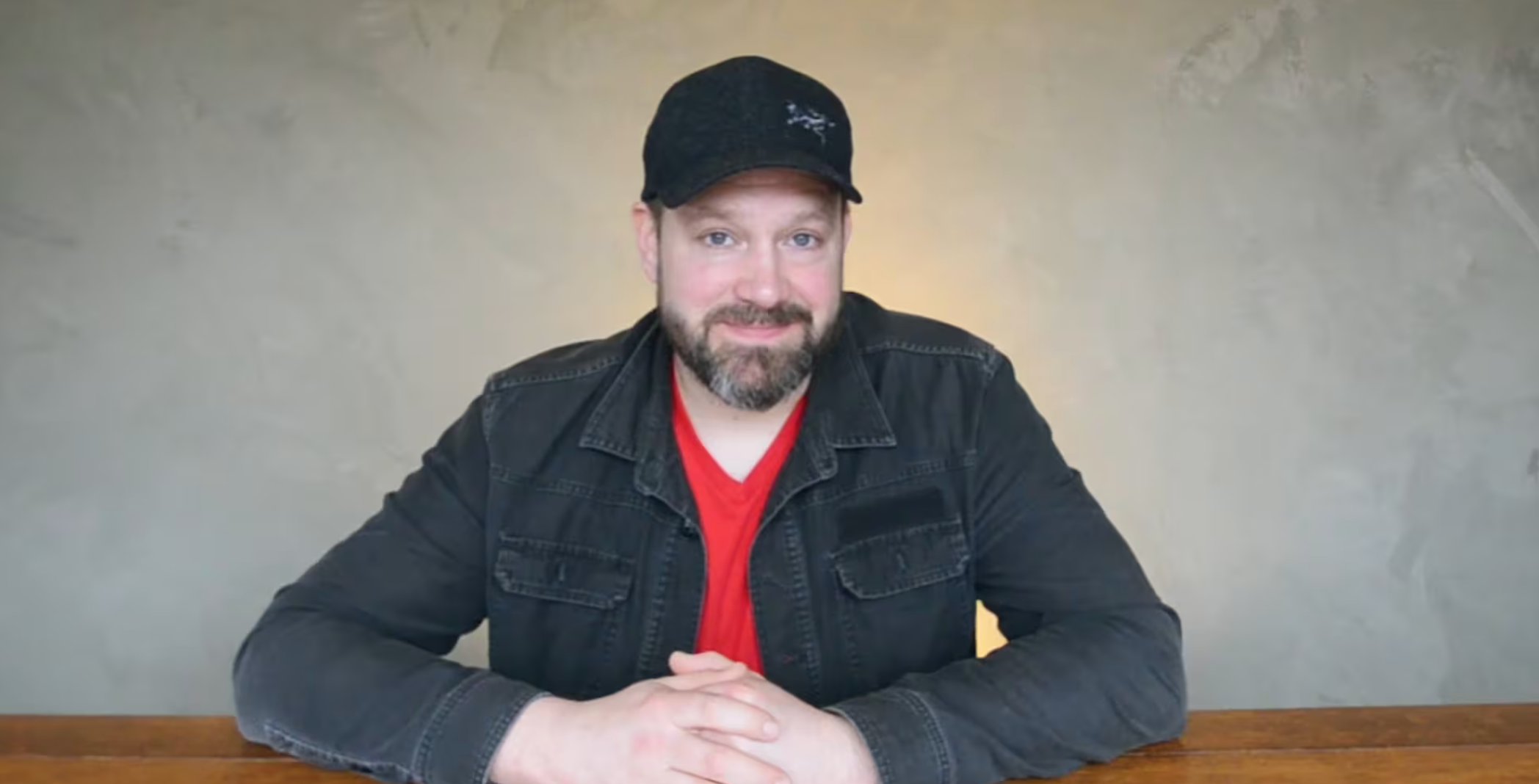Modern wooden house with large glass window, set against mountainous landscape with dry grass and small trees in foreground.