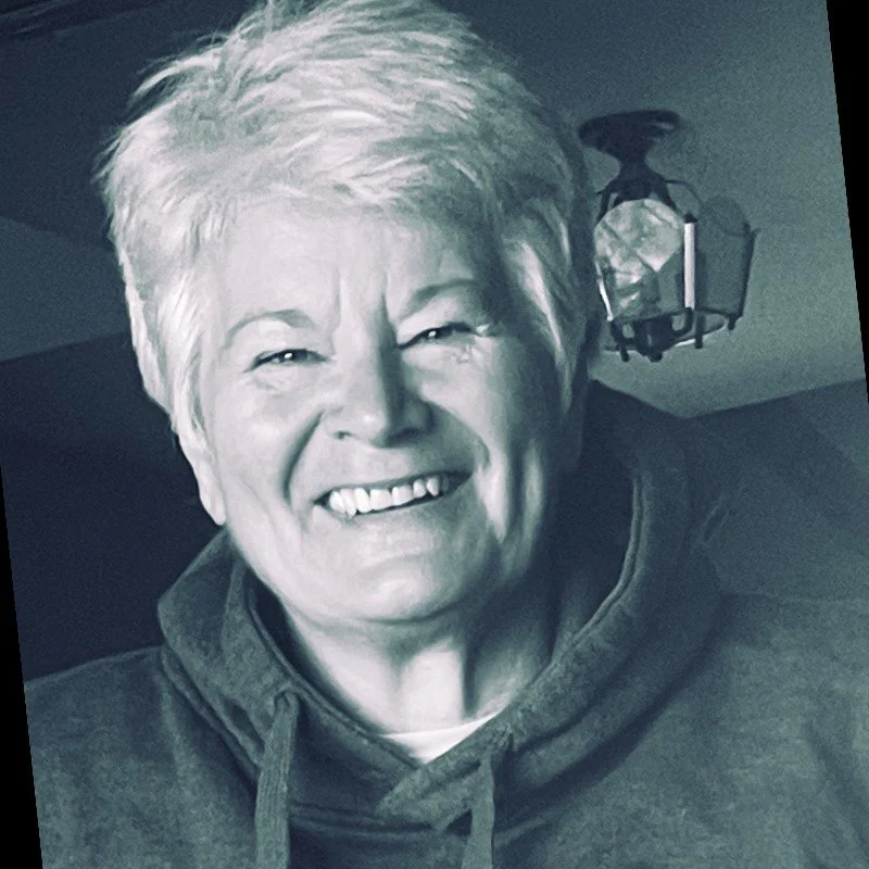 A smiling elderly woman with short, light-colored hair, wearing a dark hoodie, indoors with a ceiling light fixture in the background.