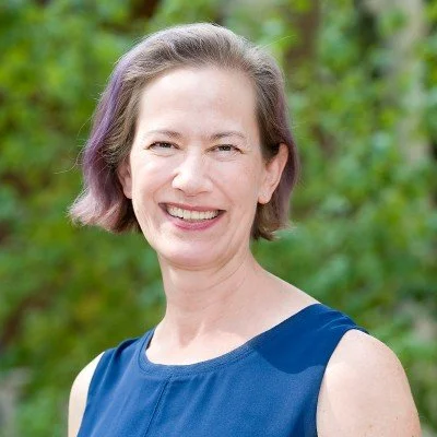 Smiling woman with short hair, wearing a blue sleeveless top, outdoors in a green, leafy setting.