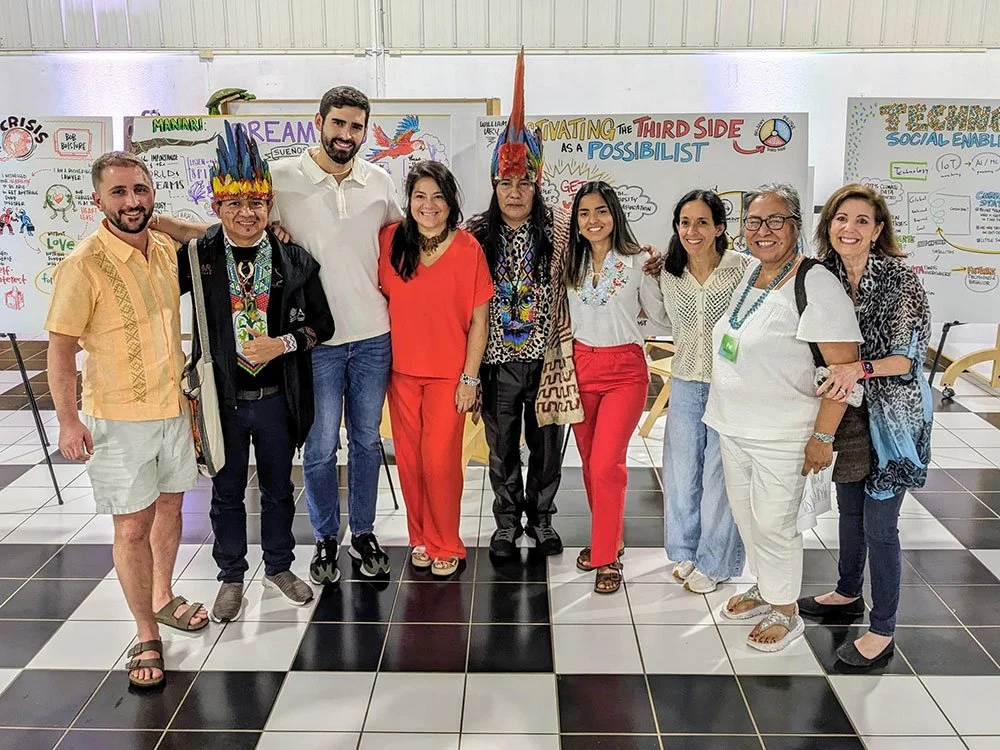 A group of nine adults standing together on a tiled floor with artwork and text on boards in the background. Two individuals are wearing colorful feather headdresses. The group is dressed in casual and traditional attire, smiling for the photo.