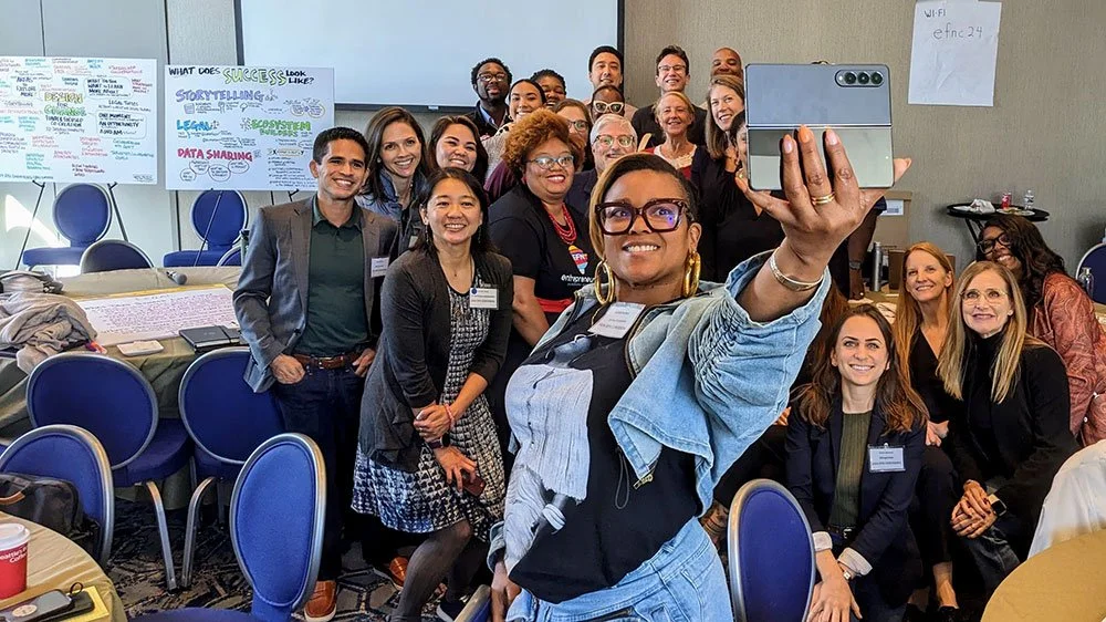 Group of people taking a selfie at a conference room, with chairs and discussion boards visible in the background.