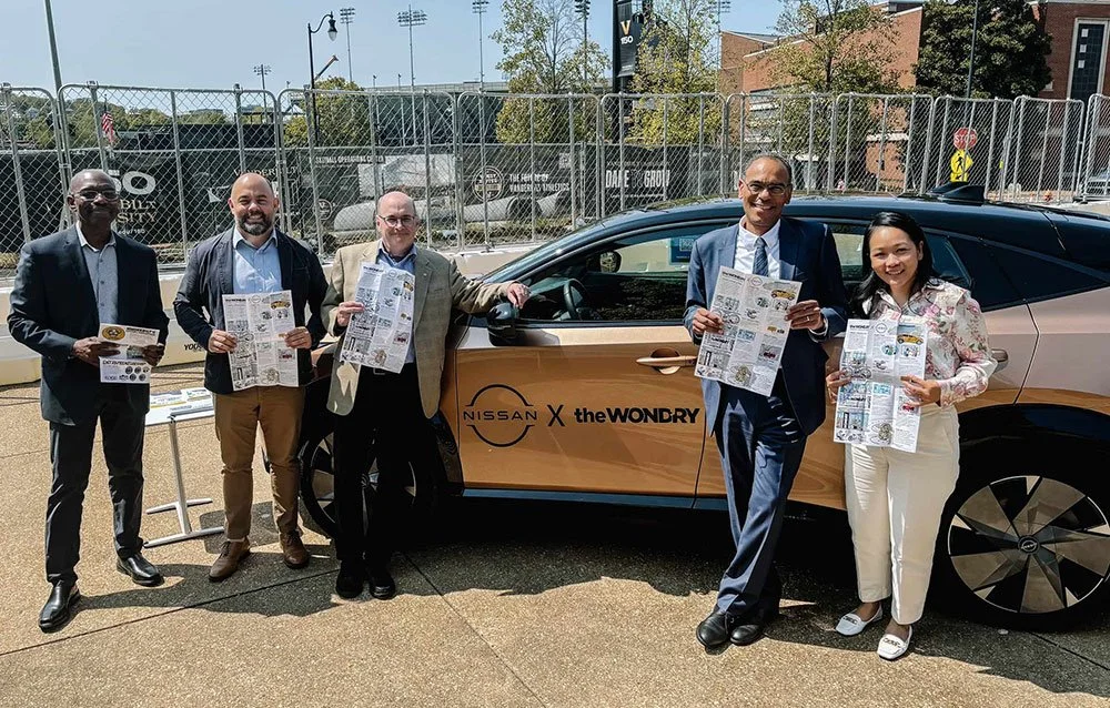 Group of five people standing next to a car with "Nissan X the Wond'ry" branding, holding brochures, outdoors.