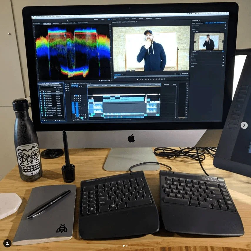An Apple iMac computer displaying video editing software with a waveform, timeline, and video preview. A person is shown on the screen holding a microphone and wearing a hoodie and cap. On the desk, there is a black water bottle with a skull design, a notebook with a pen, a small black pen holder, two ergonomic keyboards, and a graphics tablet with a stylus.