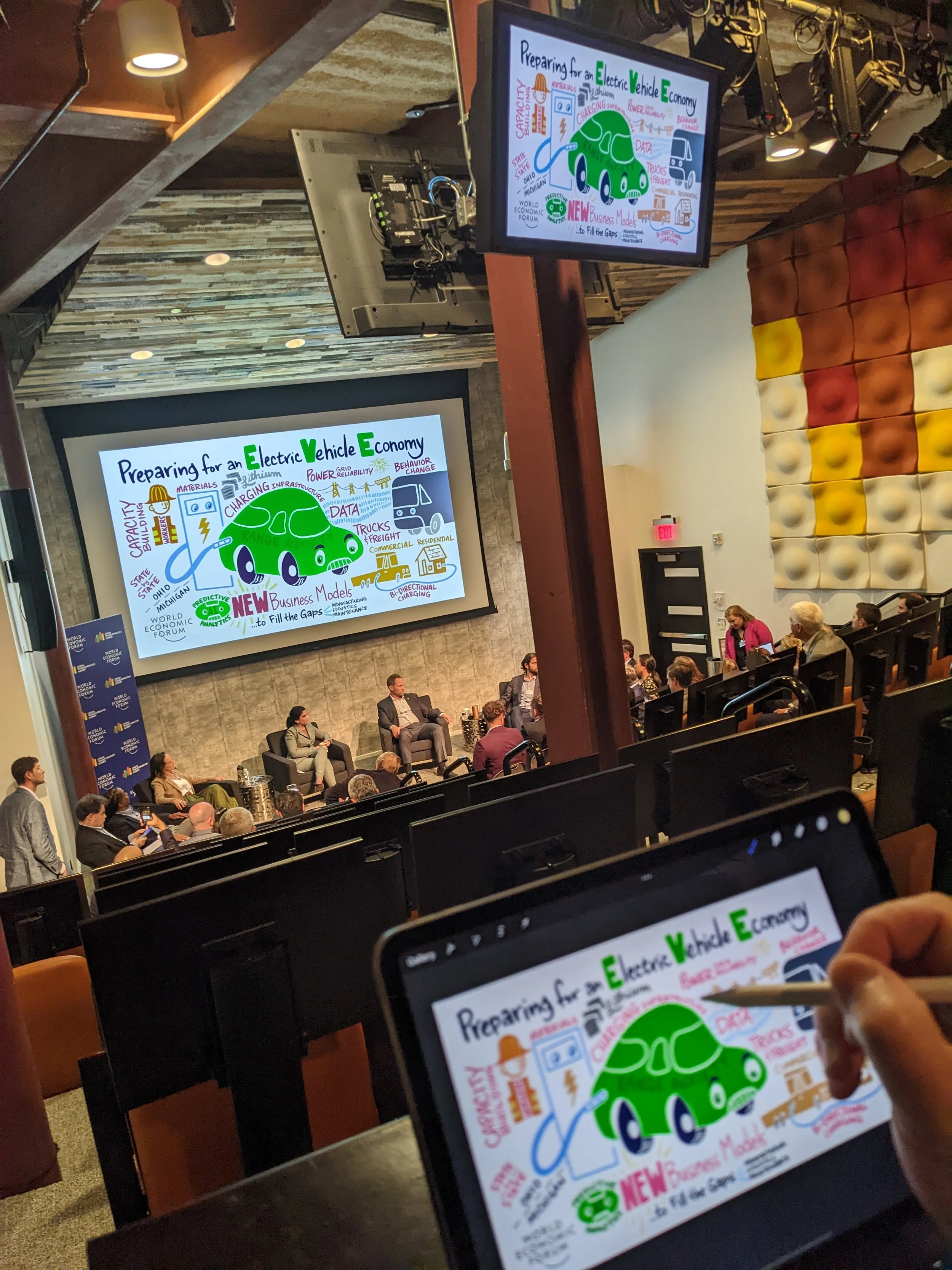 A conference room with a large screen displaying a colorful presentation slide about preparing for an electric vehicle economy. The slide features a green electric car and handwritten notes and diagrams. There are several people seated and a person taking notes on a tablet, with some attendees on stage participating in a panel discussion.