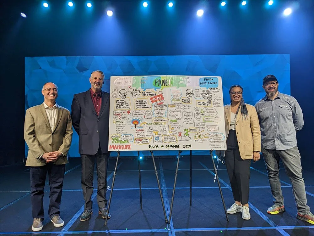 Four people standing on a stage beside a detailed, colorful illustrated poster. The blue background is illuminated by stage lights. Each person is smiling and dressed in smart casual attire.