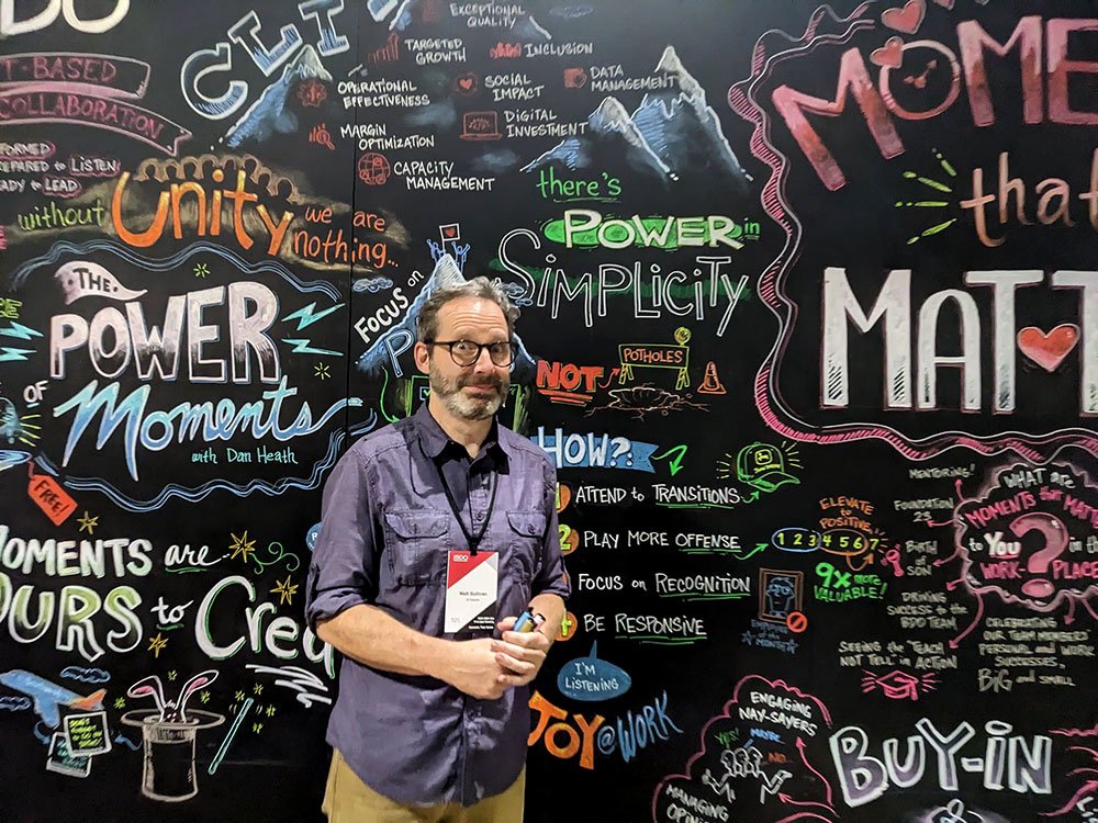 Matt Sullivan is standing in front of a colorful chalkboard wall filled with motivational phrases and drawings, such as "The Power of Moments," "Unity we are nothing," and "There's Power in Simplicity." The person holds markers and wears a name tag.