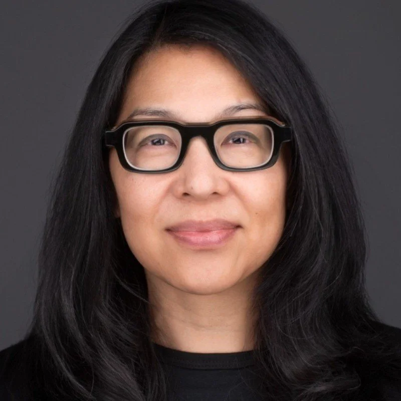 A woman with long black hair wearing black rectangular glasses smiling at the camera against a dark background.