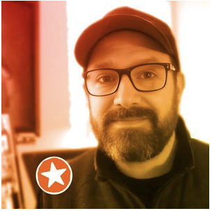 Peter Durand, glasses and beard, wearing a baseball cap, in an indoor setting with warm lighting.
