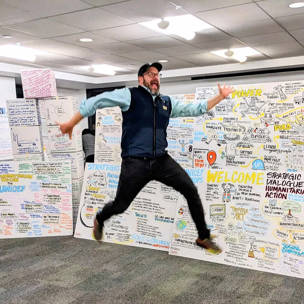 Peter Durand, founder of Alphachimp LLC,  jumping with joy in front of large colorful whiteboards covered in strategic and brainstorming notes and drawings.