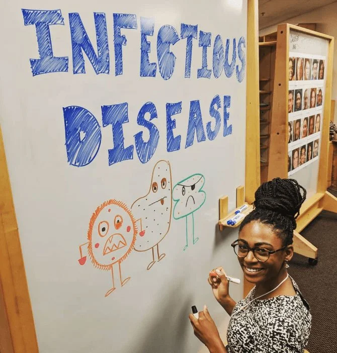 A woman with glasses and braids smiling while drawing on a whiteboard. The board has large, blue, handwritten text that reads 'INFECTIOUS DISEASE' and three cartoon-like illustrations of microorganisms in orange, brown, and teal.