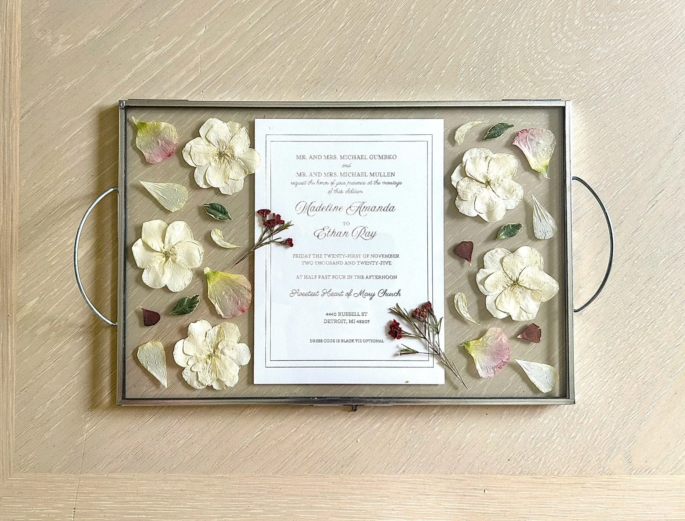 A wedding invitation displayed on a glass frame, decorated with dried flower petals and small dried flowers around the border, on a wooden surface