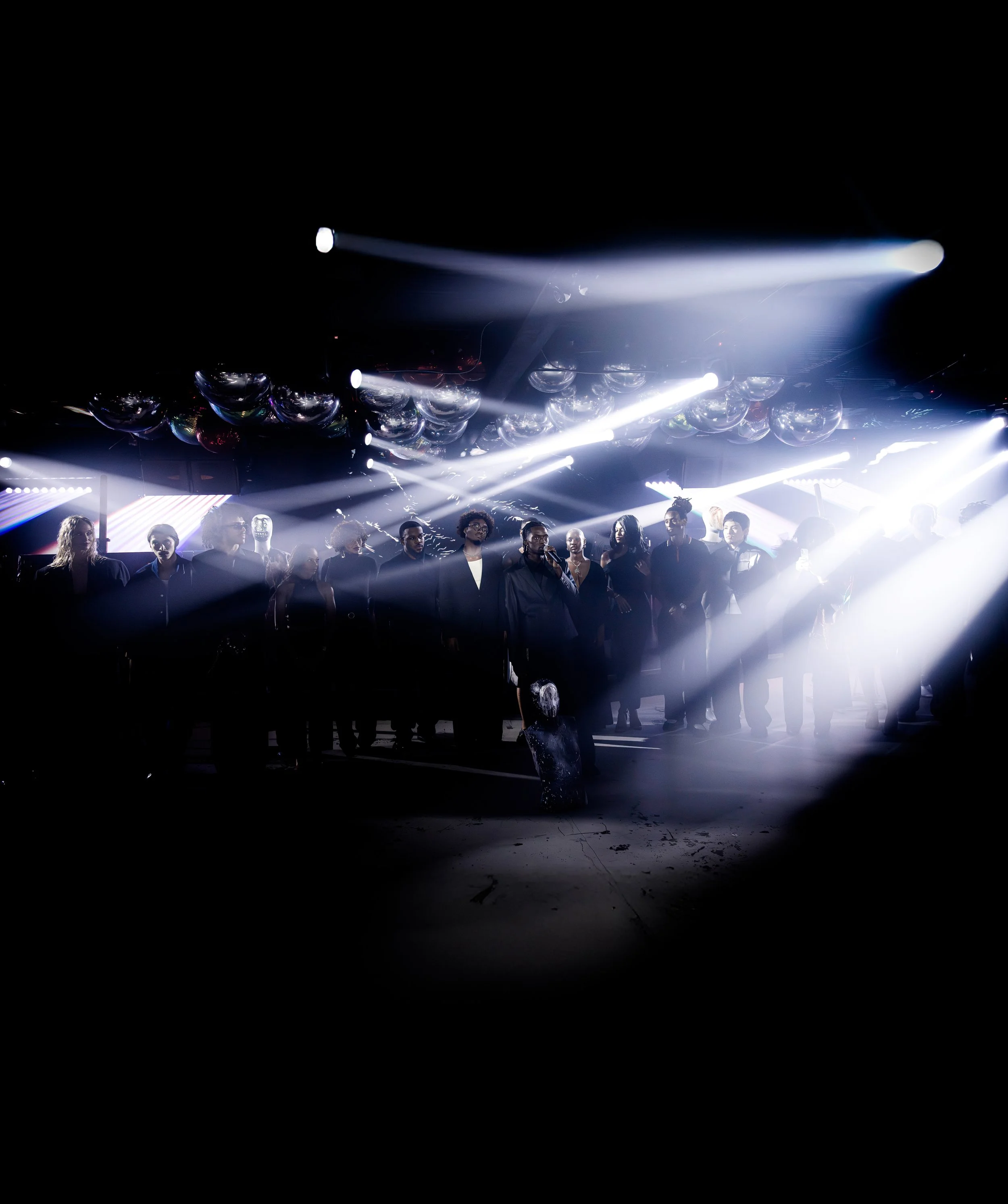 Group of people standing in a dark room illuminated by bright stage lights, with reflective balloons hanging from the ceiling.