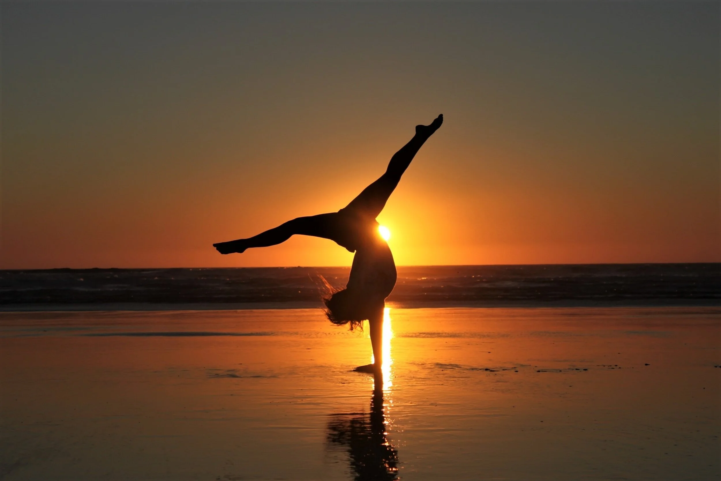 A person performing a handstand on the beach at sunset, with the sun setting behind them and their silhouette reflected in the water.