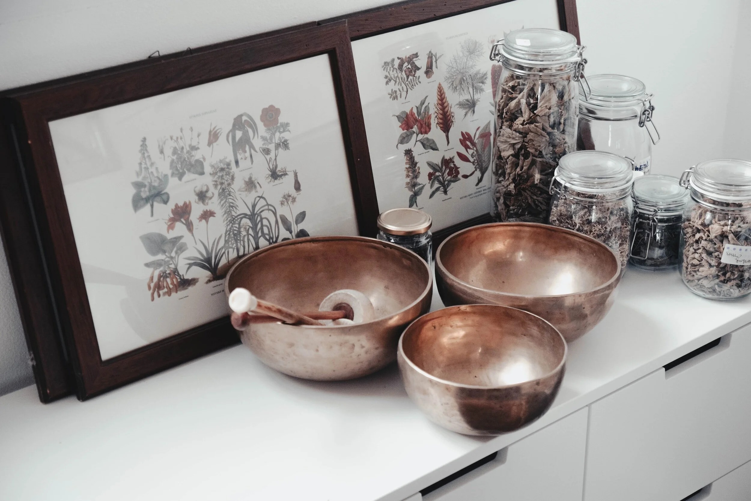 Three bronze singing bowls on a white surface, behind which are framed botanical prints and glass jars filled with dried leaves or herbs.