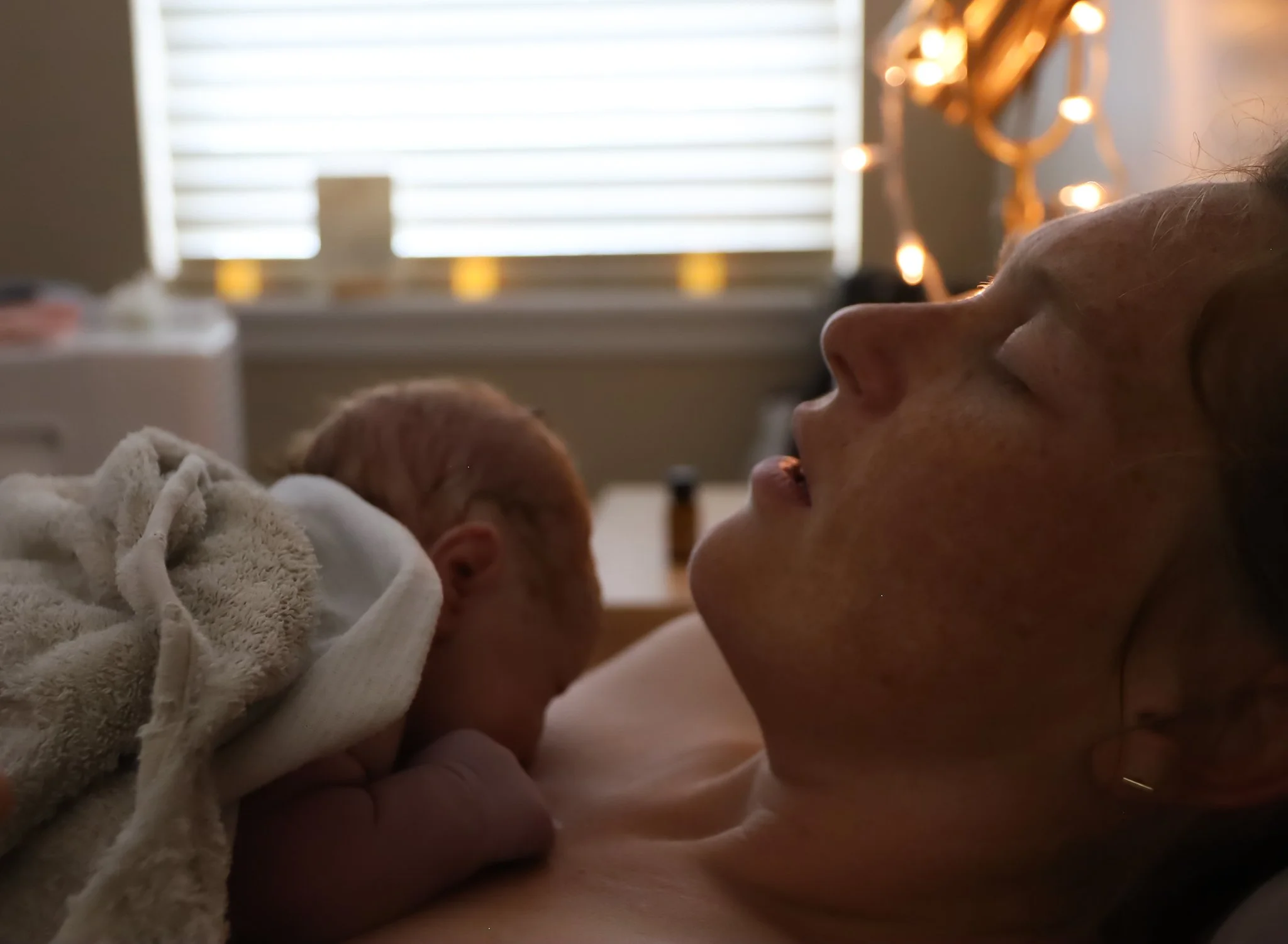 A woman lying in bed with a newborn baby resting on her chest, in a dimly lit room with sunlight filtering through blinds and string lights in the background.