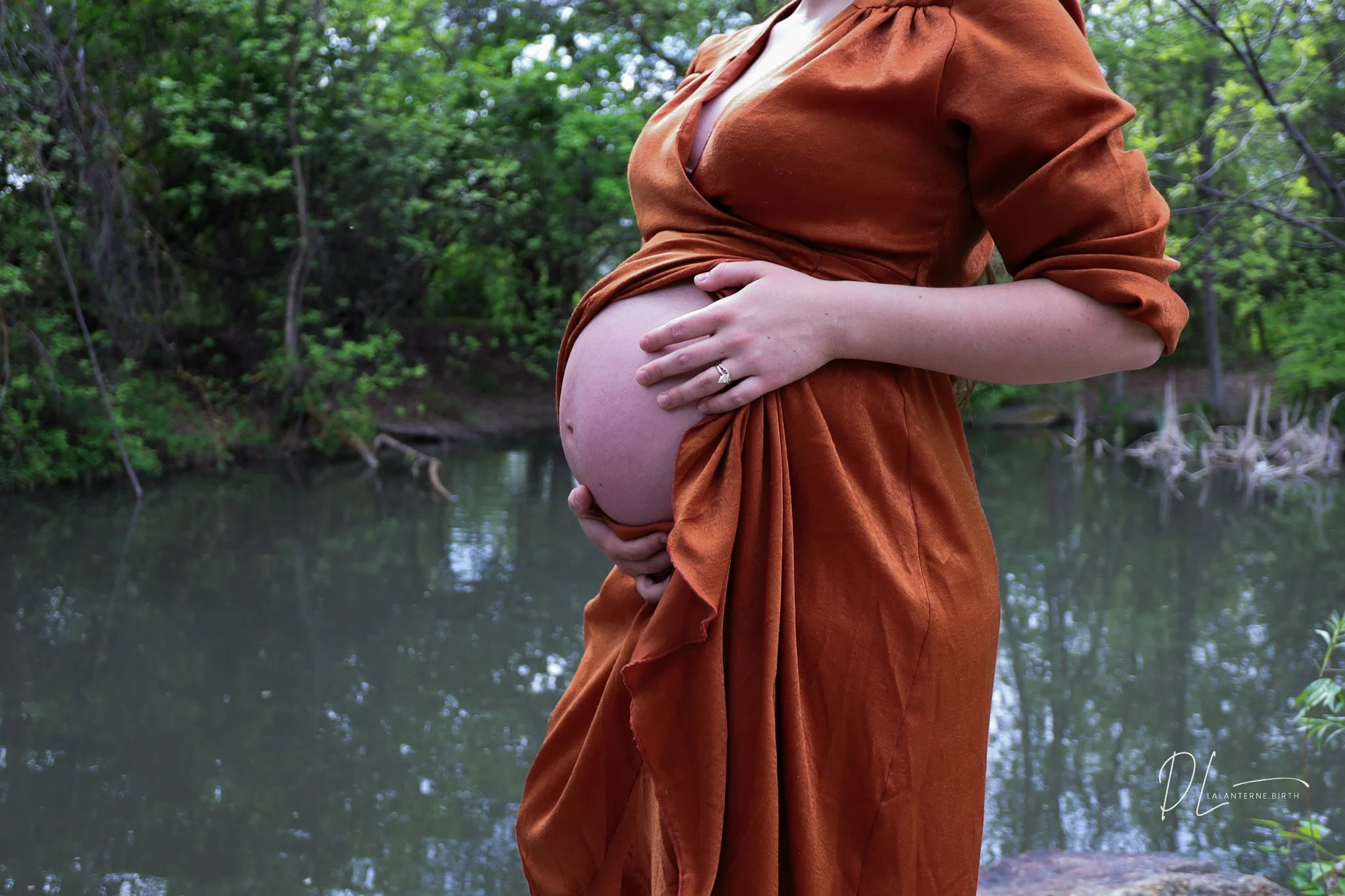 A pregnant woman wearing a rust-colored dress standing near a river in a lush green forest.