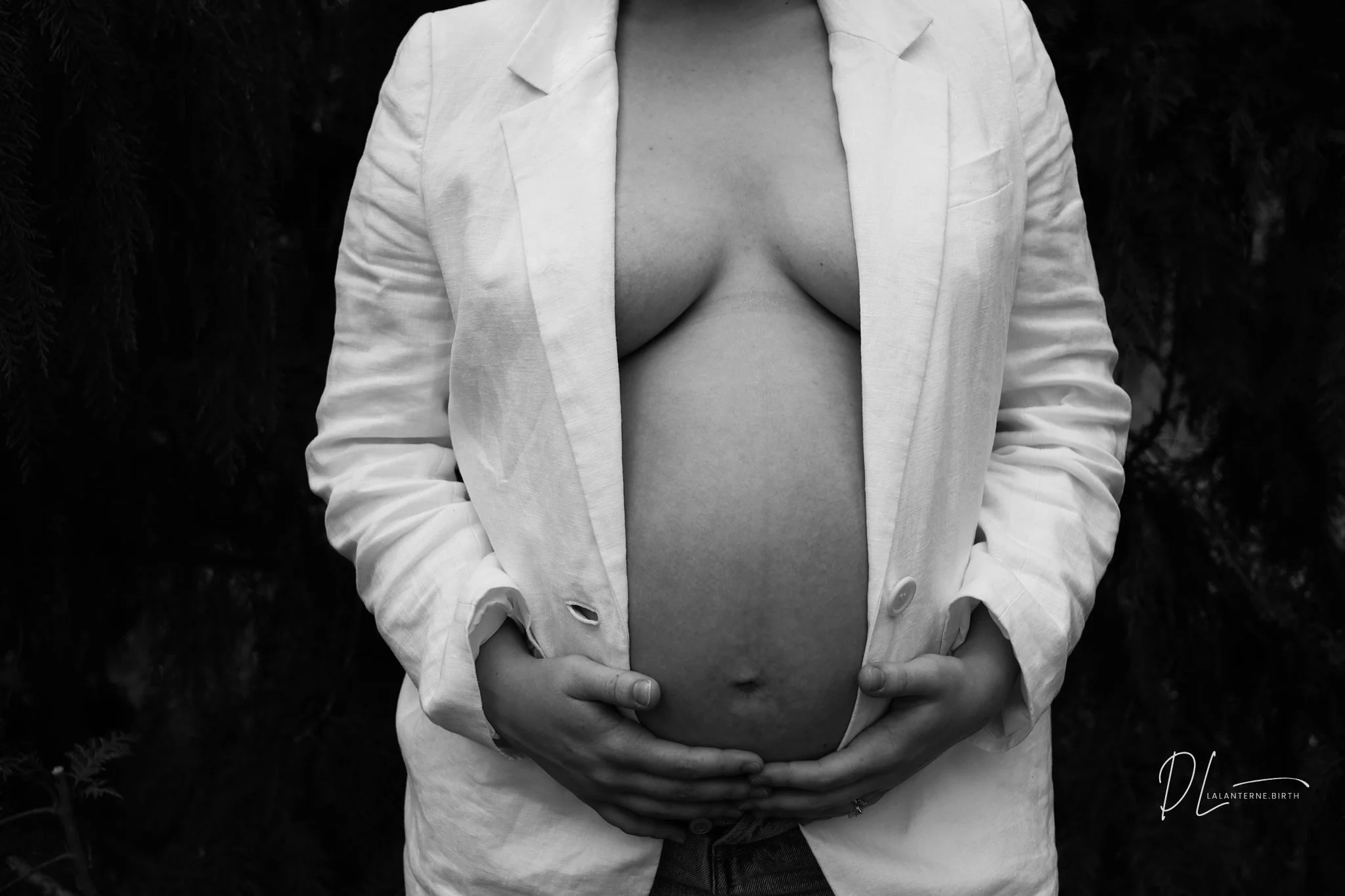 A pregnant woman with her torso exposed, wearing an open white blazer and holding her belly with her hands. The image is in black and white.