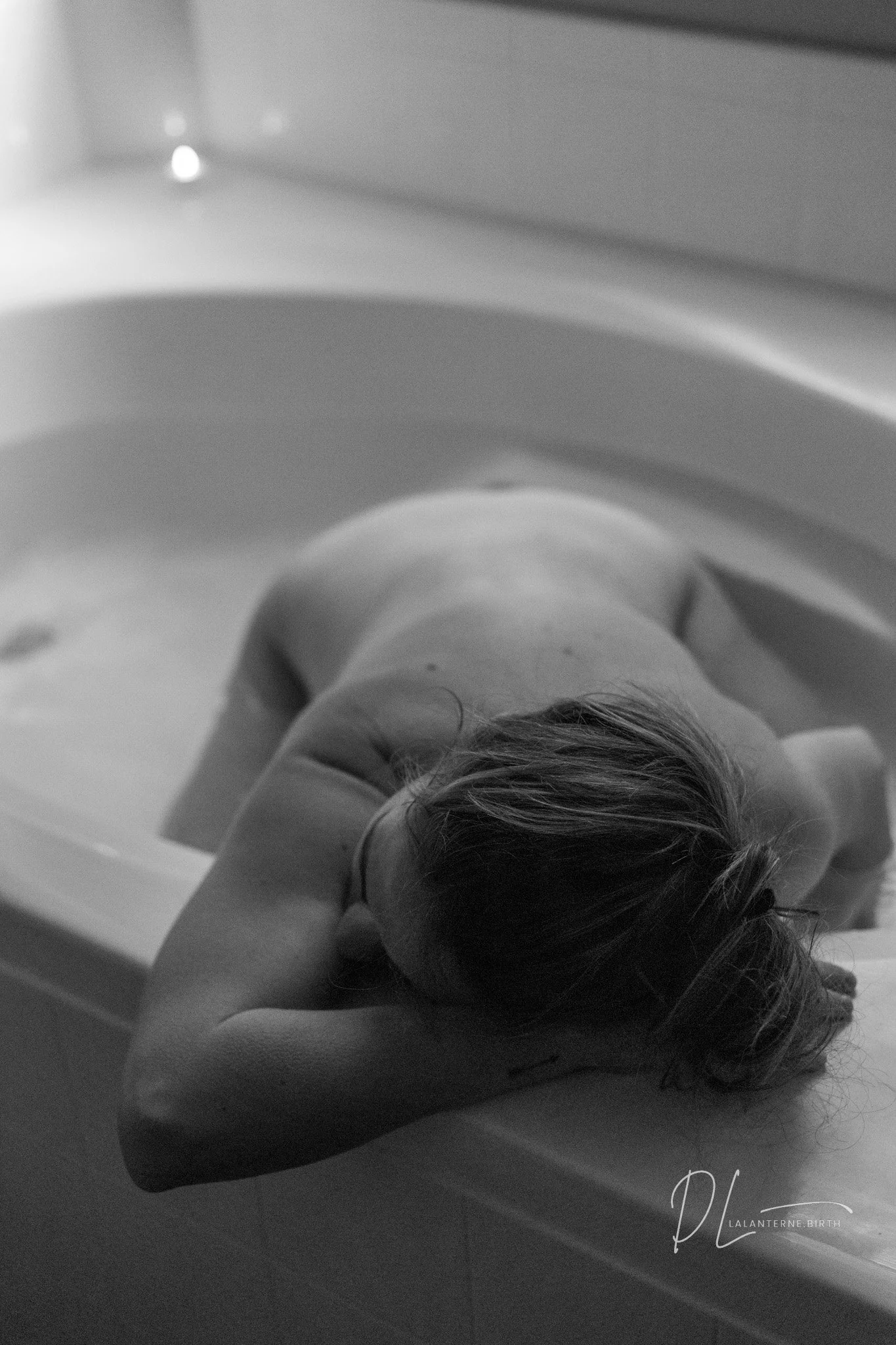 A woman lying on a bathtub edge in a bathroom, with her head resting on her arms, captured in black and white.
