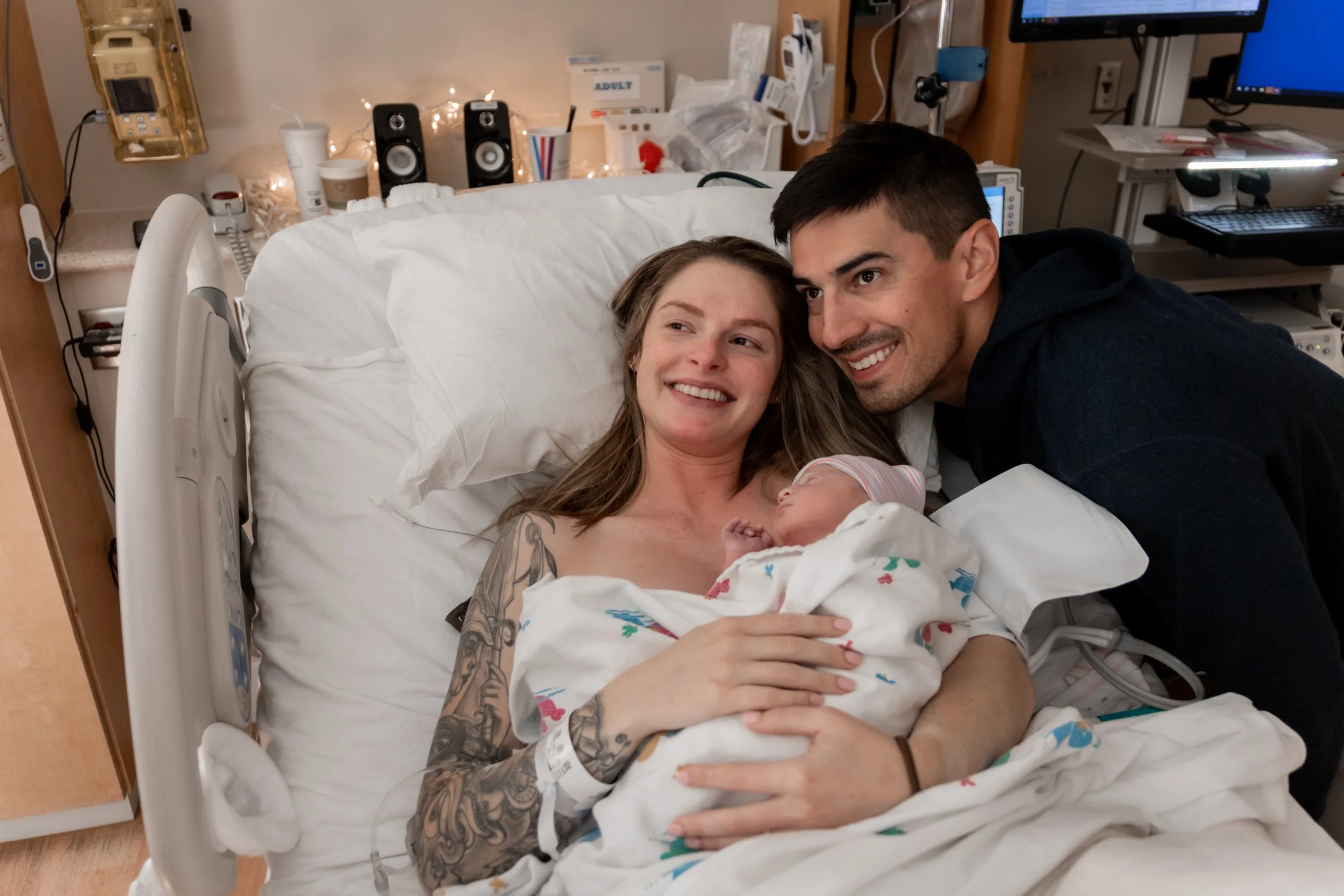 A mother holding a newborn in a hospital bed with a man leaning over her, all smiling.