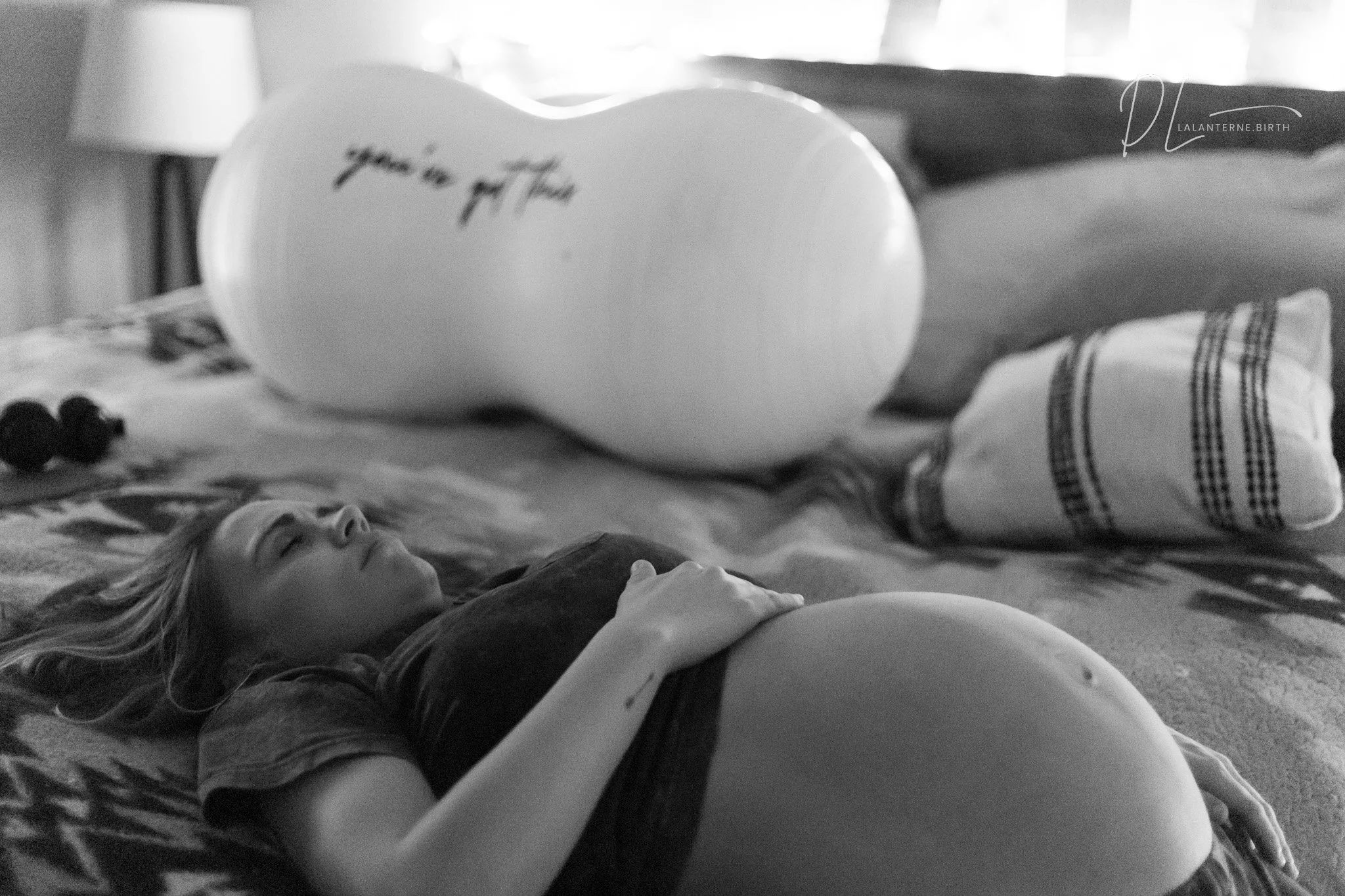A woman lying on a bed, holding her pregnant belly, with a large balloon on the bed that says 'you're in my thoughts', and a smaller pillow with a striped pattern beside her.