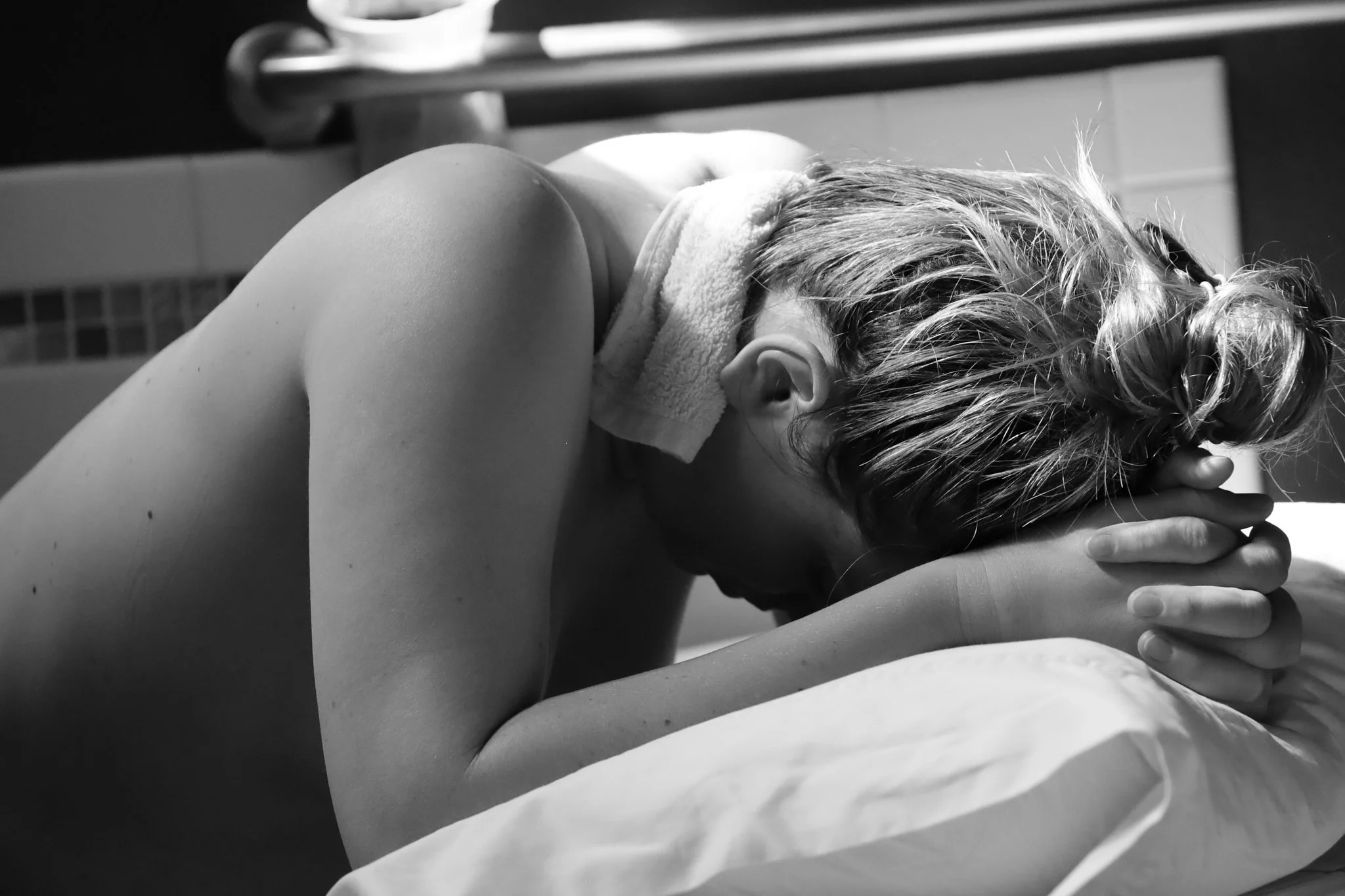 A woman with short hair, resting her head on her folded arms on a pillow, appears to be in distress or deep thought in a black-and-white photo.