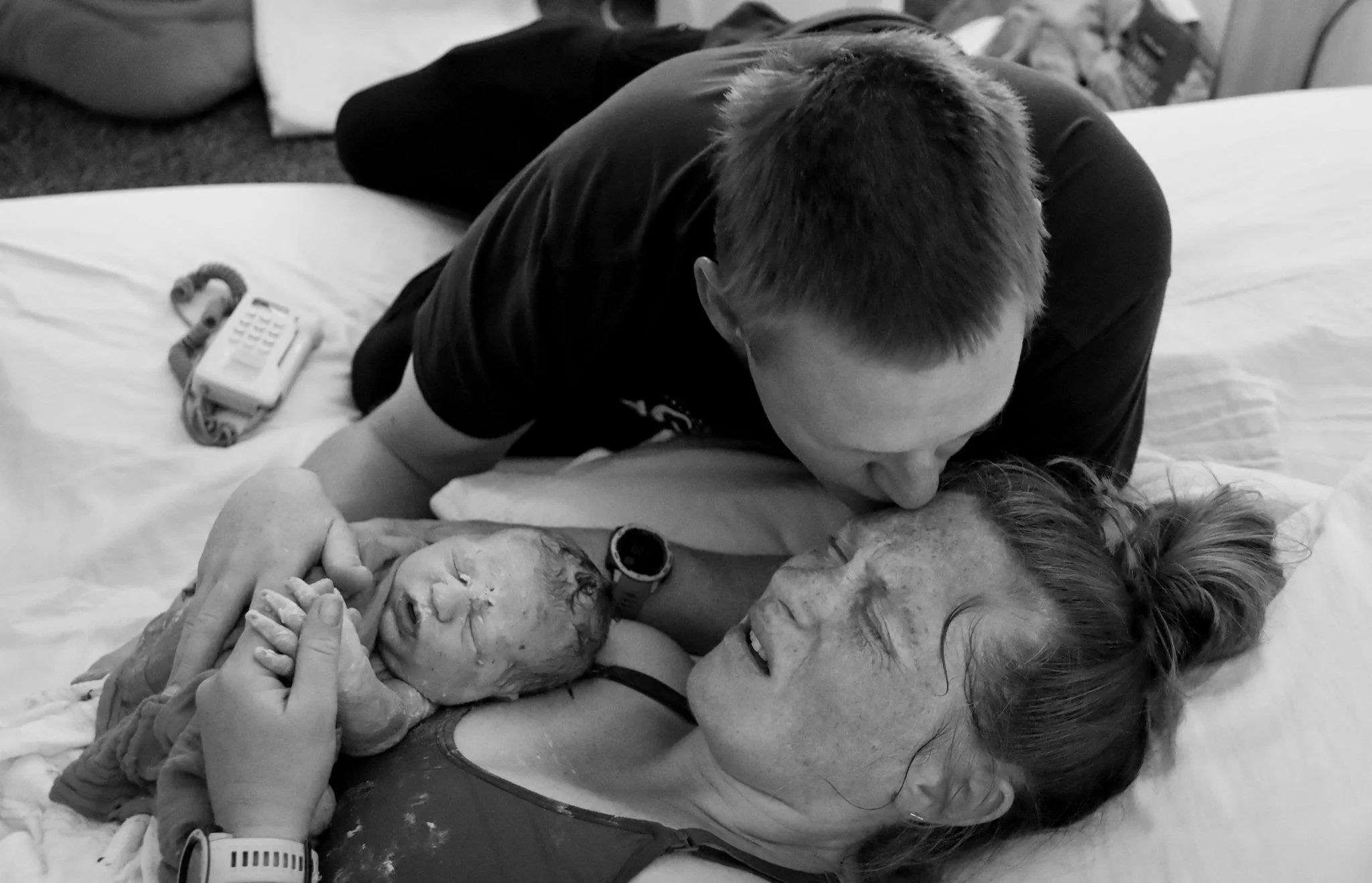 A woman lying on a bed holding a newborn baby, with a man leaning over them kissing the baby's forehead in a hospital room.