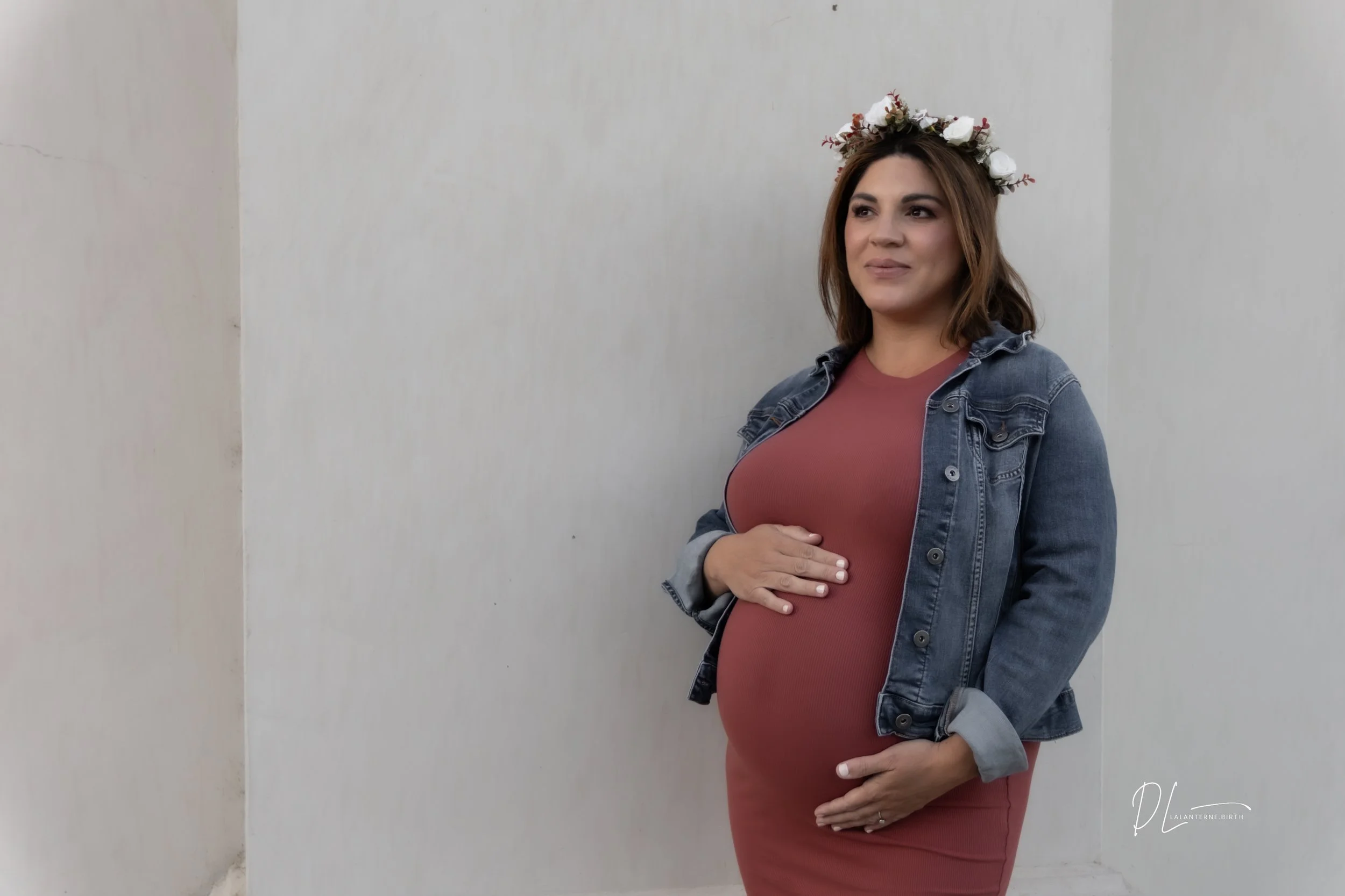 A pregnant woman wearing a denim jacket and a pink dress, with a flower crown, standing against a plain wall, gently holding her belly.