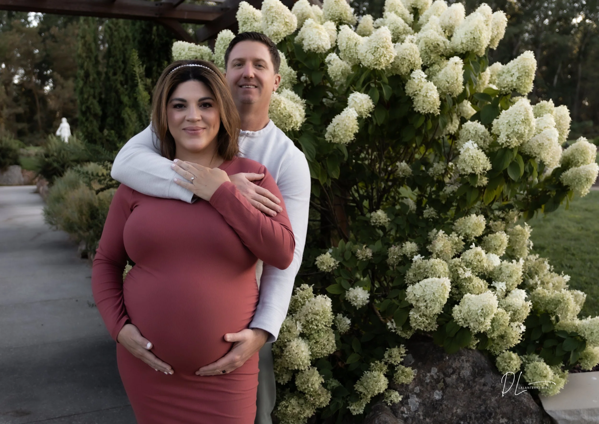 A pregnant woman in a pink dress and a man in a white shirt stand outdoors, with the man hugging her from behind. They are standing next to a large flowering bush with white blossoms, in a garden setting.