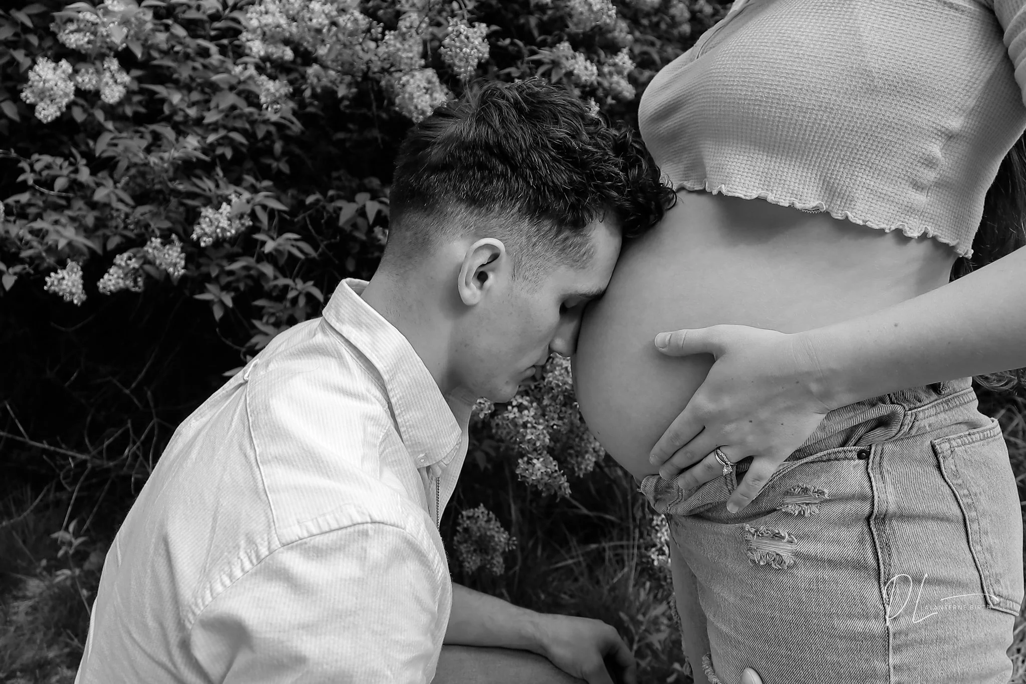 A man kneeling and touching a pregnant woman's belly with his face resting on it during an outdoor photo shoot.