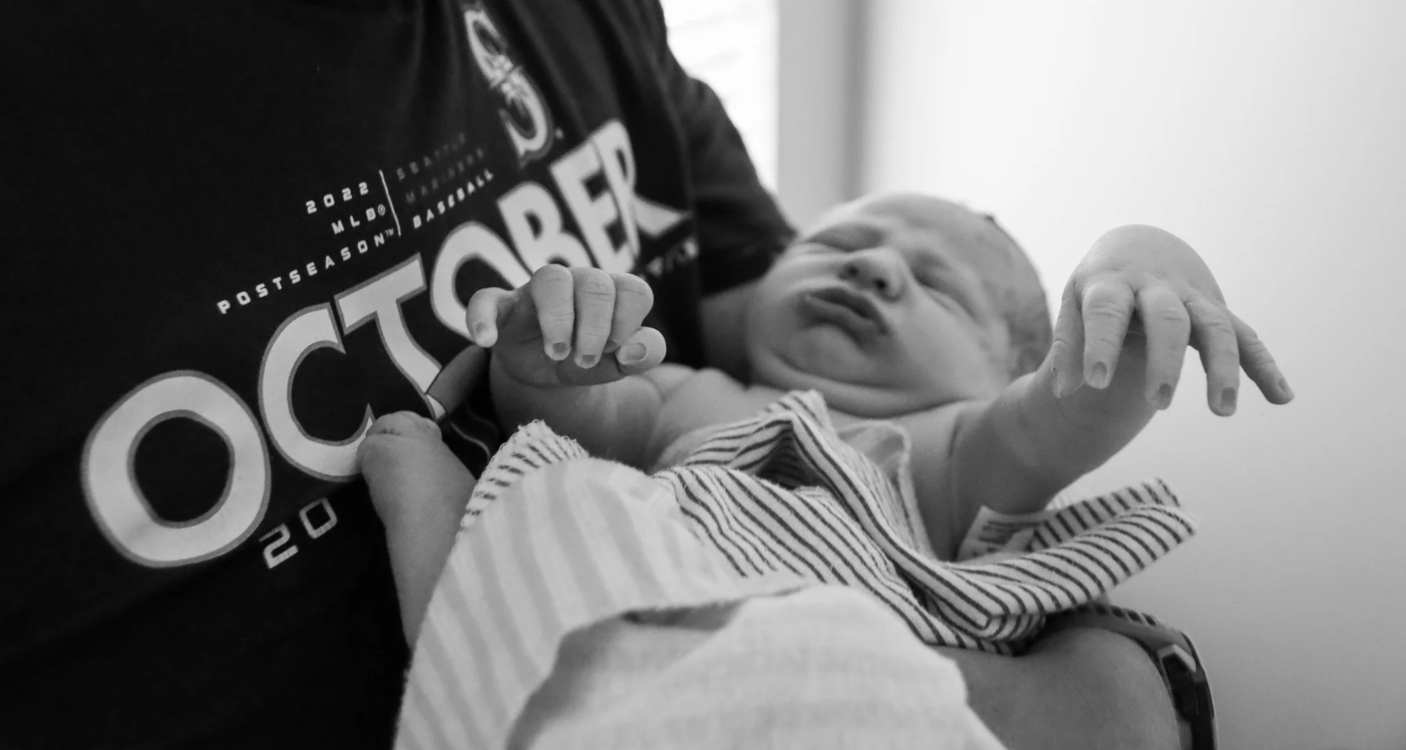 A person wearing a shirt with baseball logos is holding a newborn baby. The baby appears to be in a relaxed or sleepy state, dressed in a striped outfit, with its hand gently resting on the person holding it.