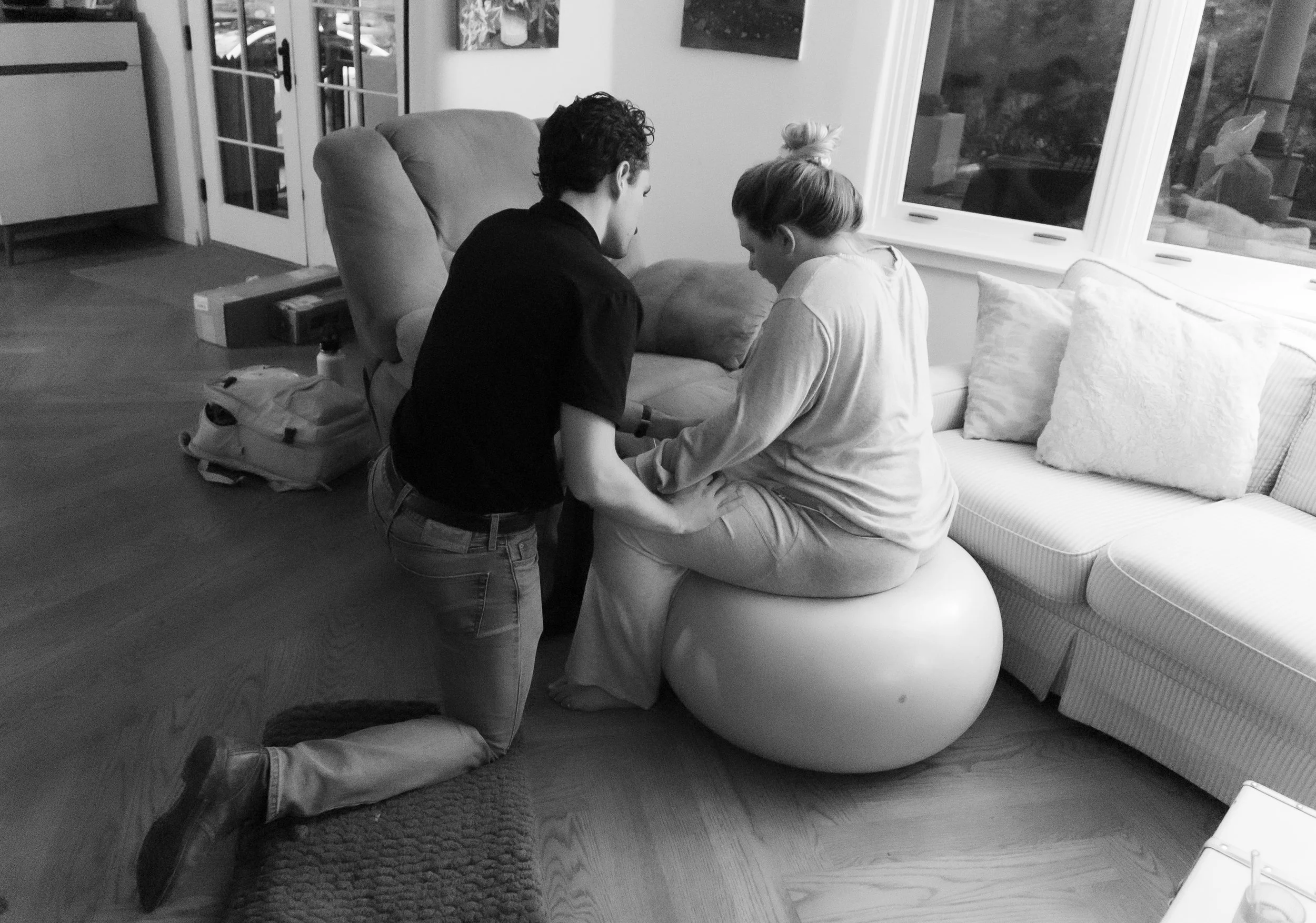 A woman sitting on an exercise ball in a living room, with a man kneeling in front of her, holding her hands. The room has a couch, cushions, a window, and various items on the floor.