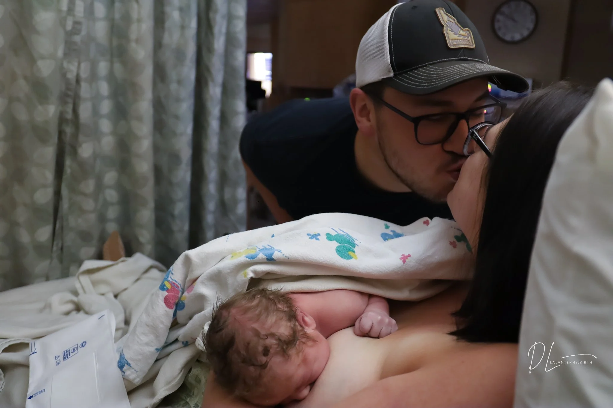 Newborn baby nursing while parents share a kiss in a hospital room.