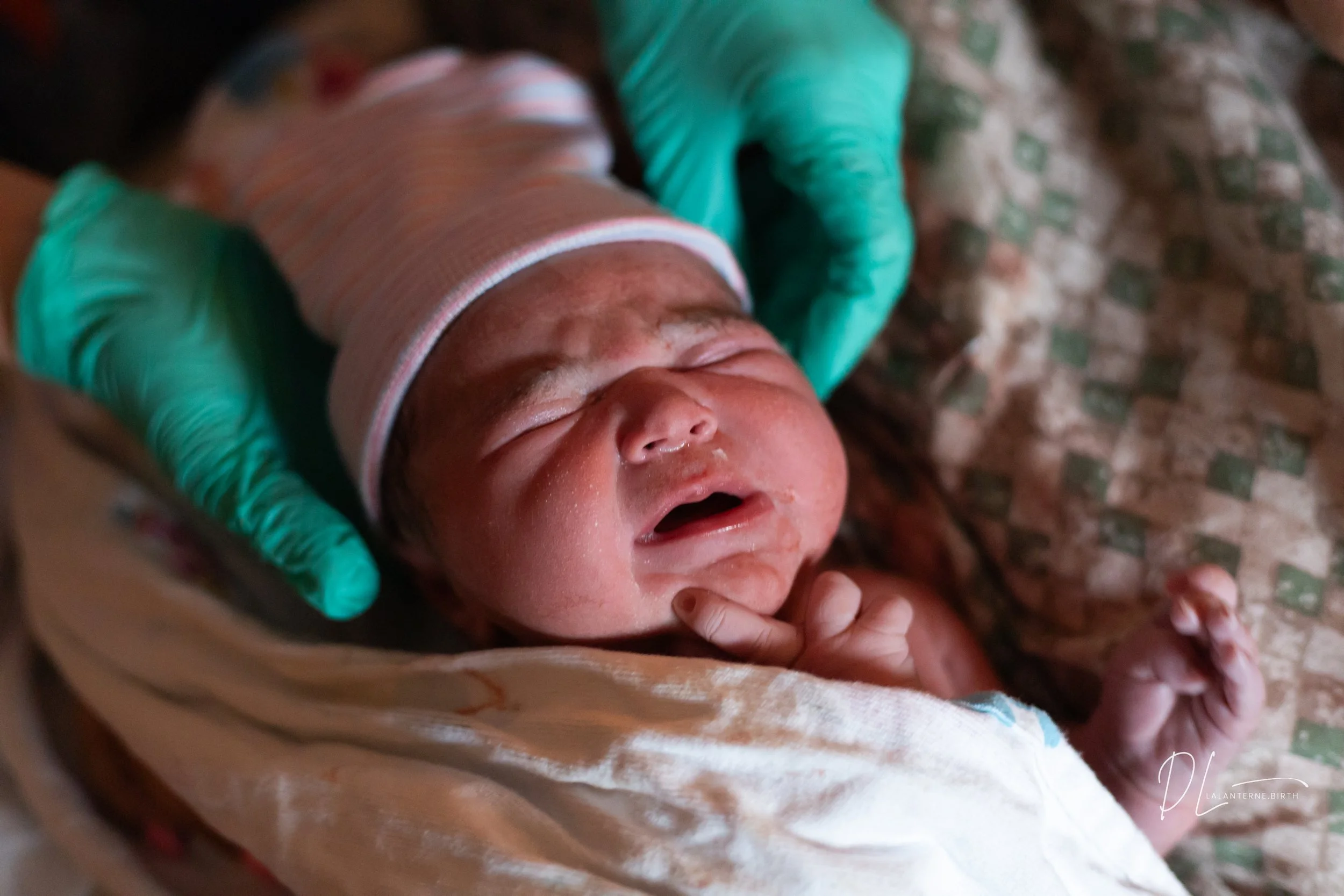 Newborn baby being held by medical professional wearing green gloves, wrapped in a cloth with a pink-striped hat, appears to be crying or in distress.