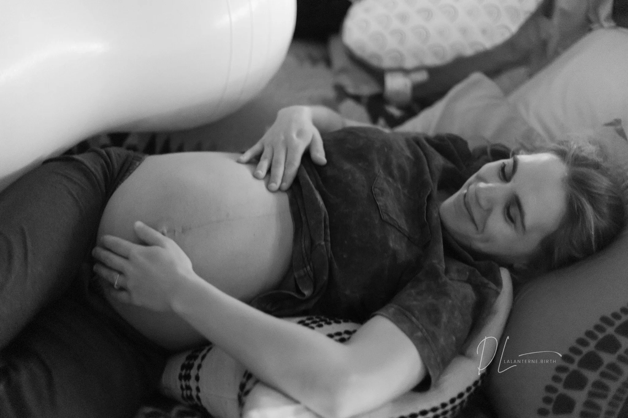 A pregnant woman lying on her side on a bed, smiling and touching her belly, with her hand resting on the top of her belly.