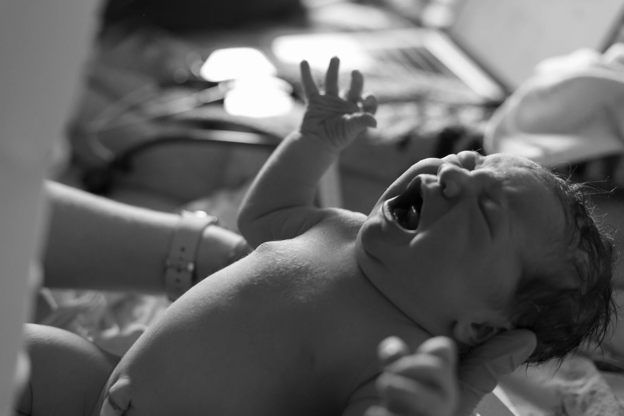 A newborn baby lying on its back, crying with eyes closed, mouth open, and hand raised. A medical professional's hand supports the baby's head.