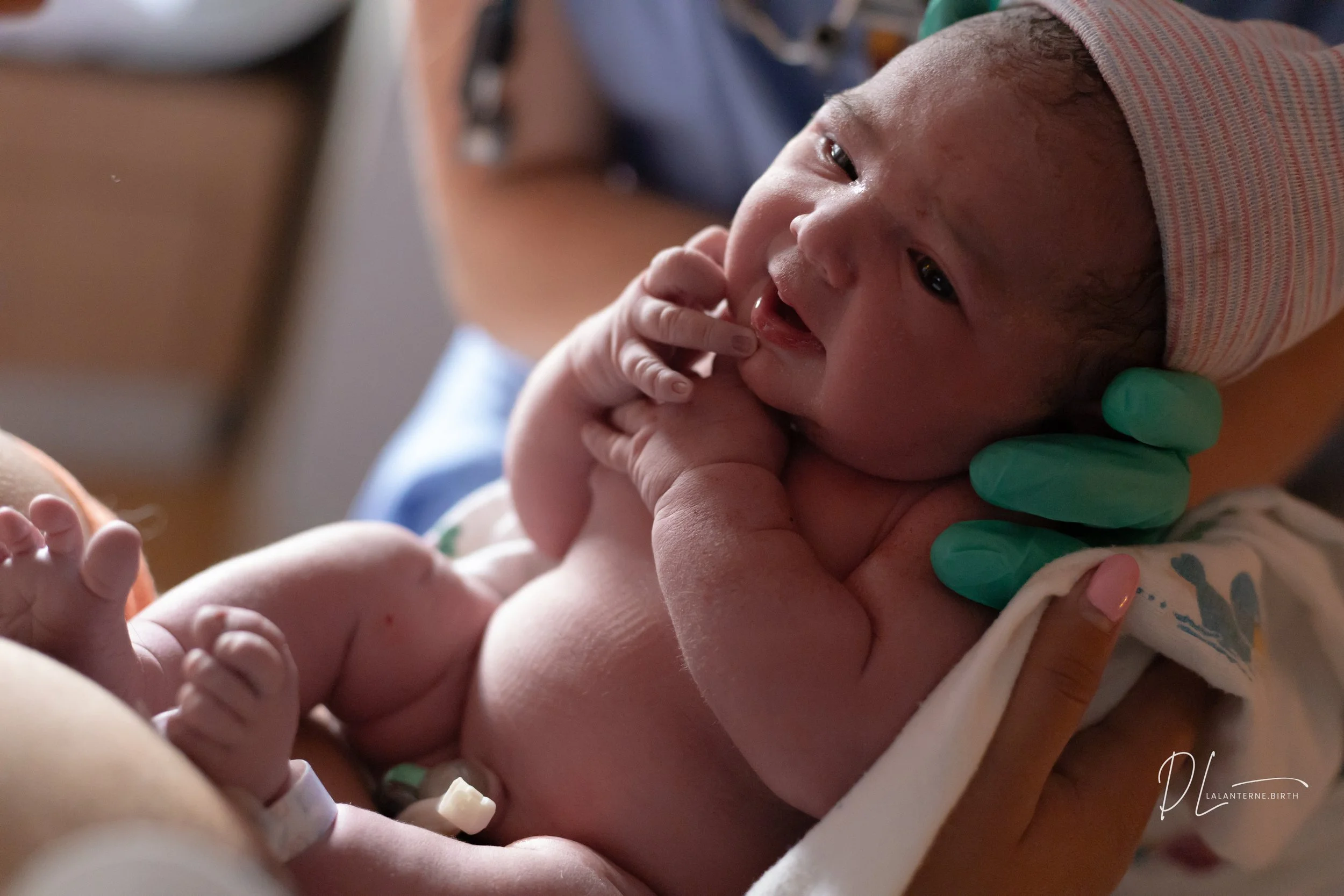 Newborn baby being held by medical staff in a hospital, with the baby’s mouth open and hand near face, wearing a striped cap.