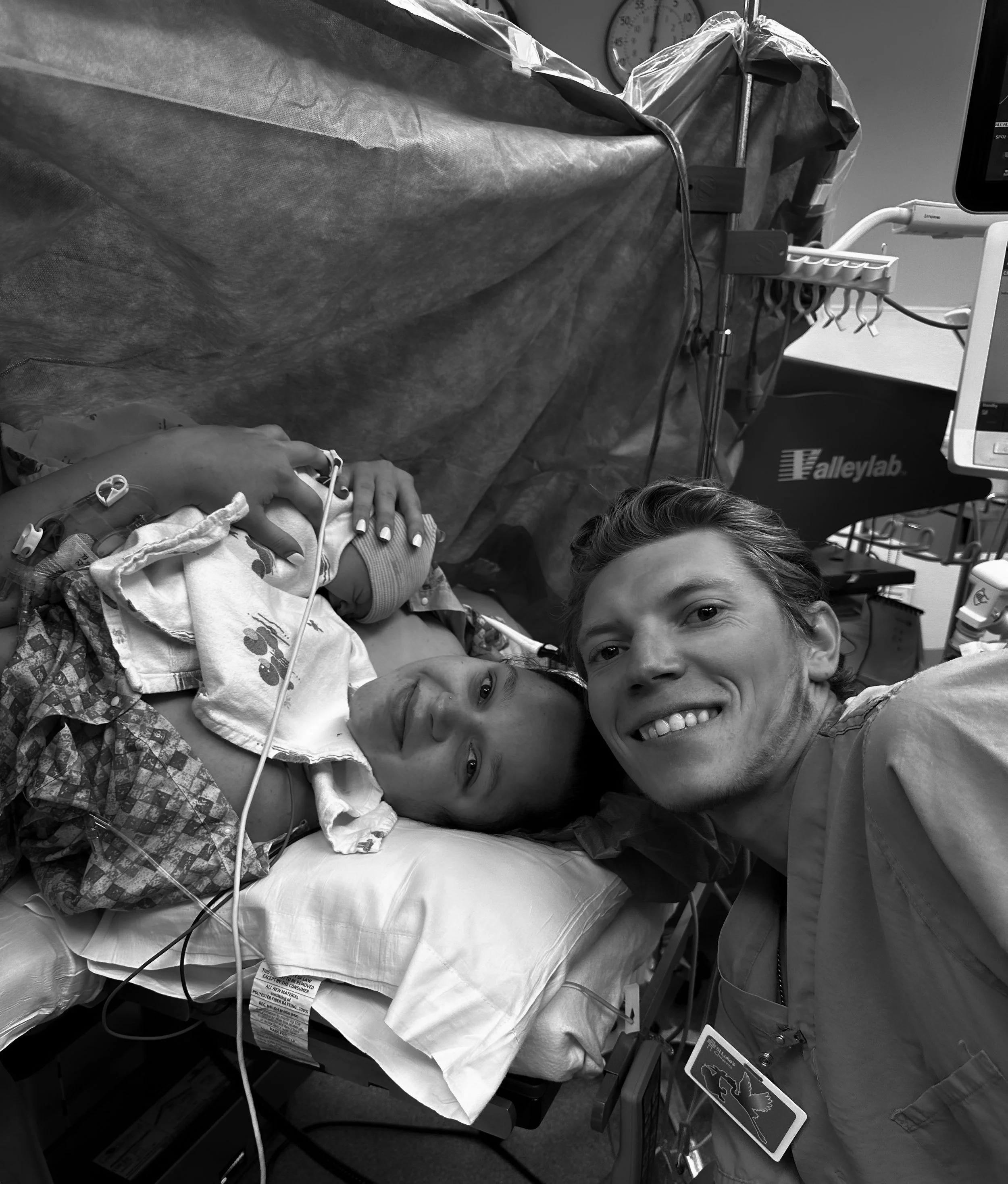 A young woman lying in a hospital bed with medical tubes attached, smiling and holding hands with a man taking a selfie beside her, in a hospital room.
