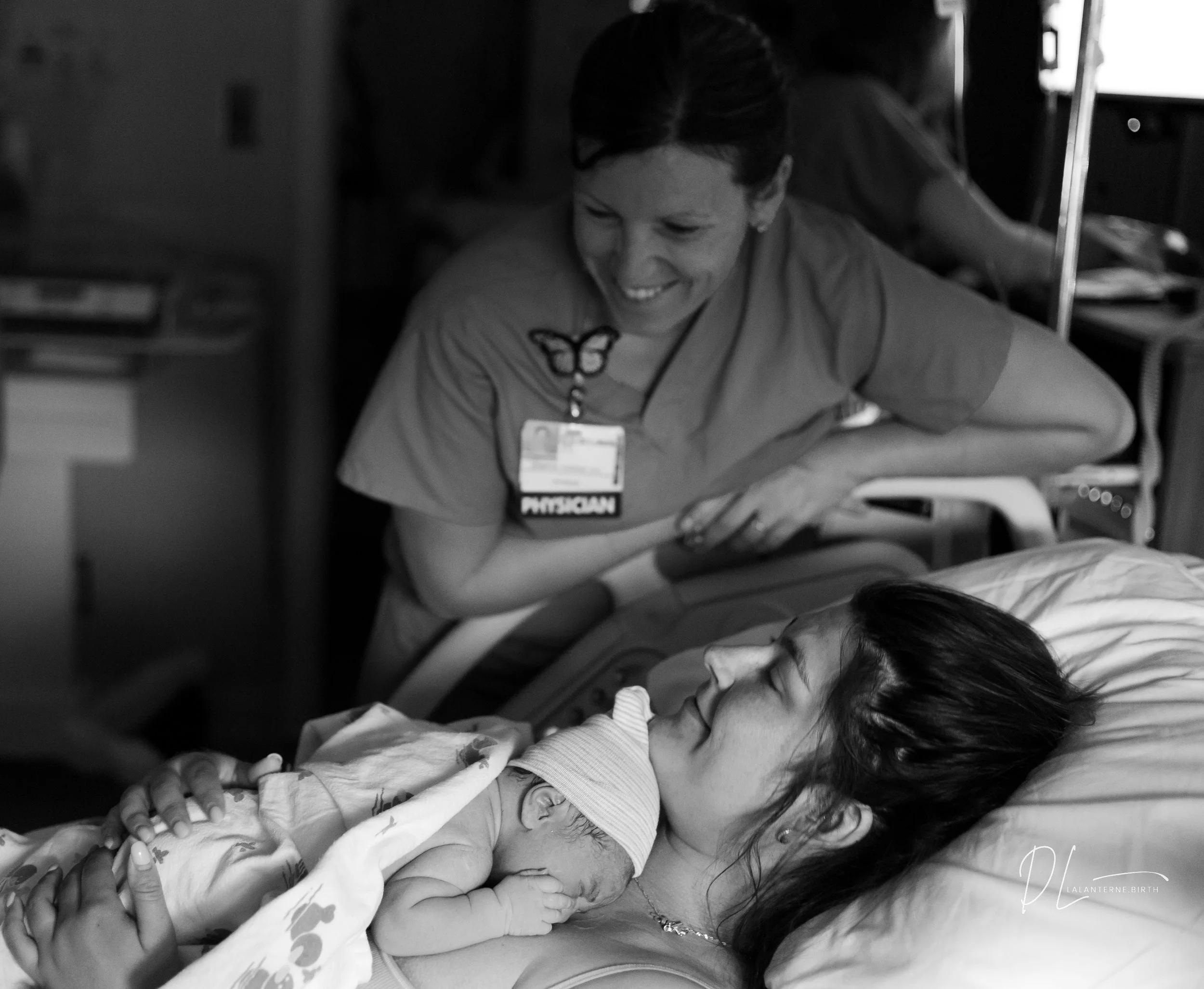 A woman resting in a hospital bed holding a newborn baby on her chest, with a smiling healthcare worker leaning over her, looking at the baby and woman.