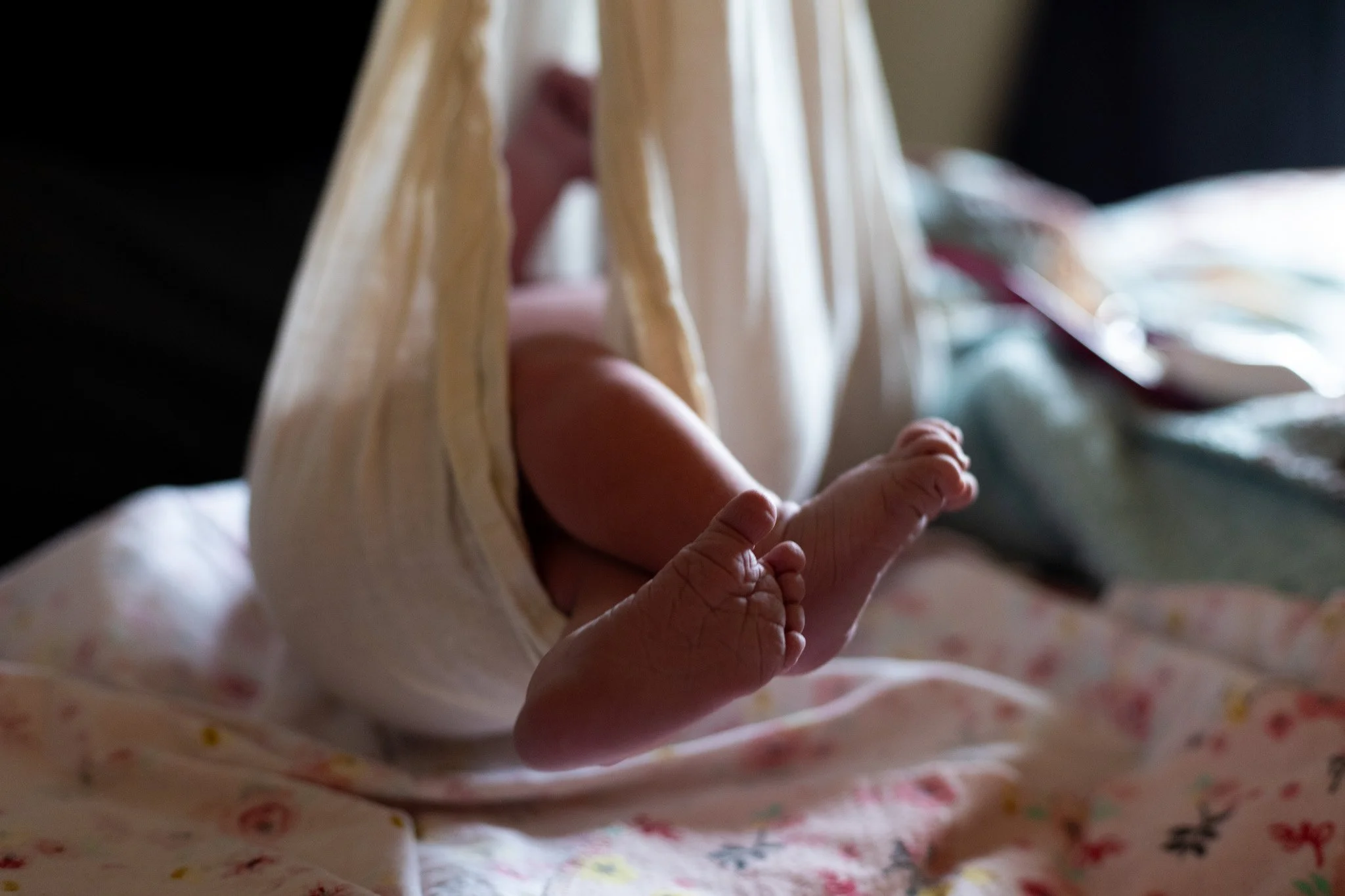 A baby is lying on their stomach on a bed, with their legs up and feet near their face. The baby is partially supported by a pillow under their chest.