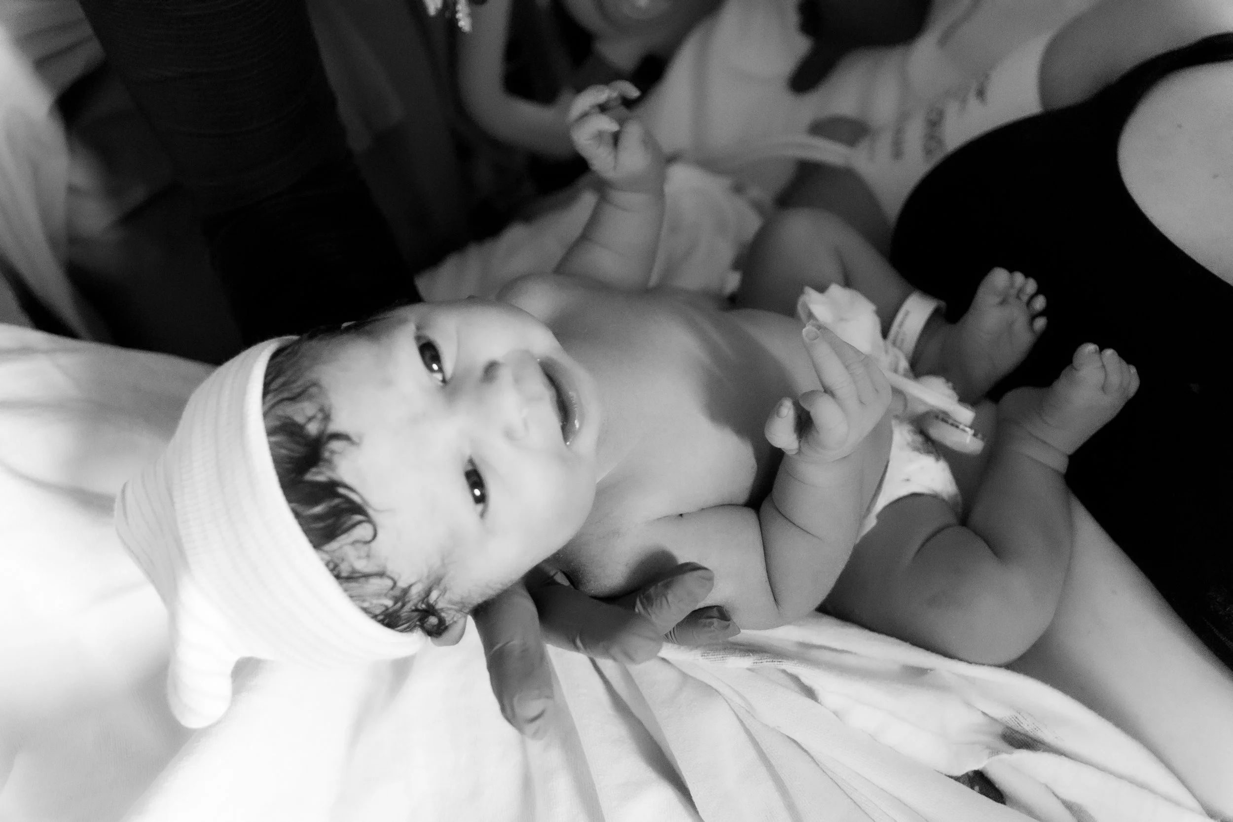 A smiling newborn baby in a hospital setting, holding a tissue in one hand, with a caregiver's hand supporting the baby.