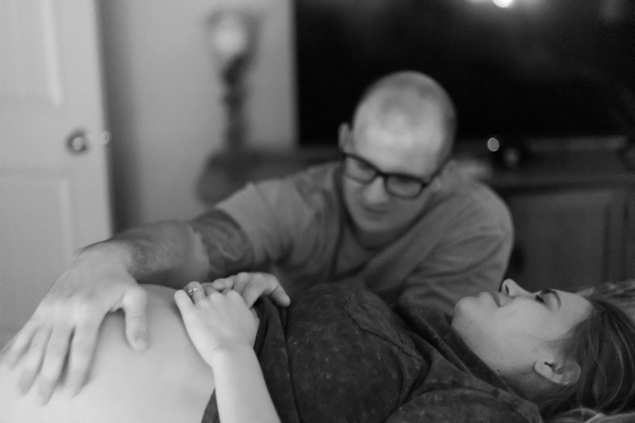 A person lying on a bed with a woman sitting beside them, holding their hand and touching their stomach. The man is smiling and wearing glasses. The woman is looking at the man and smiling.