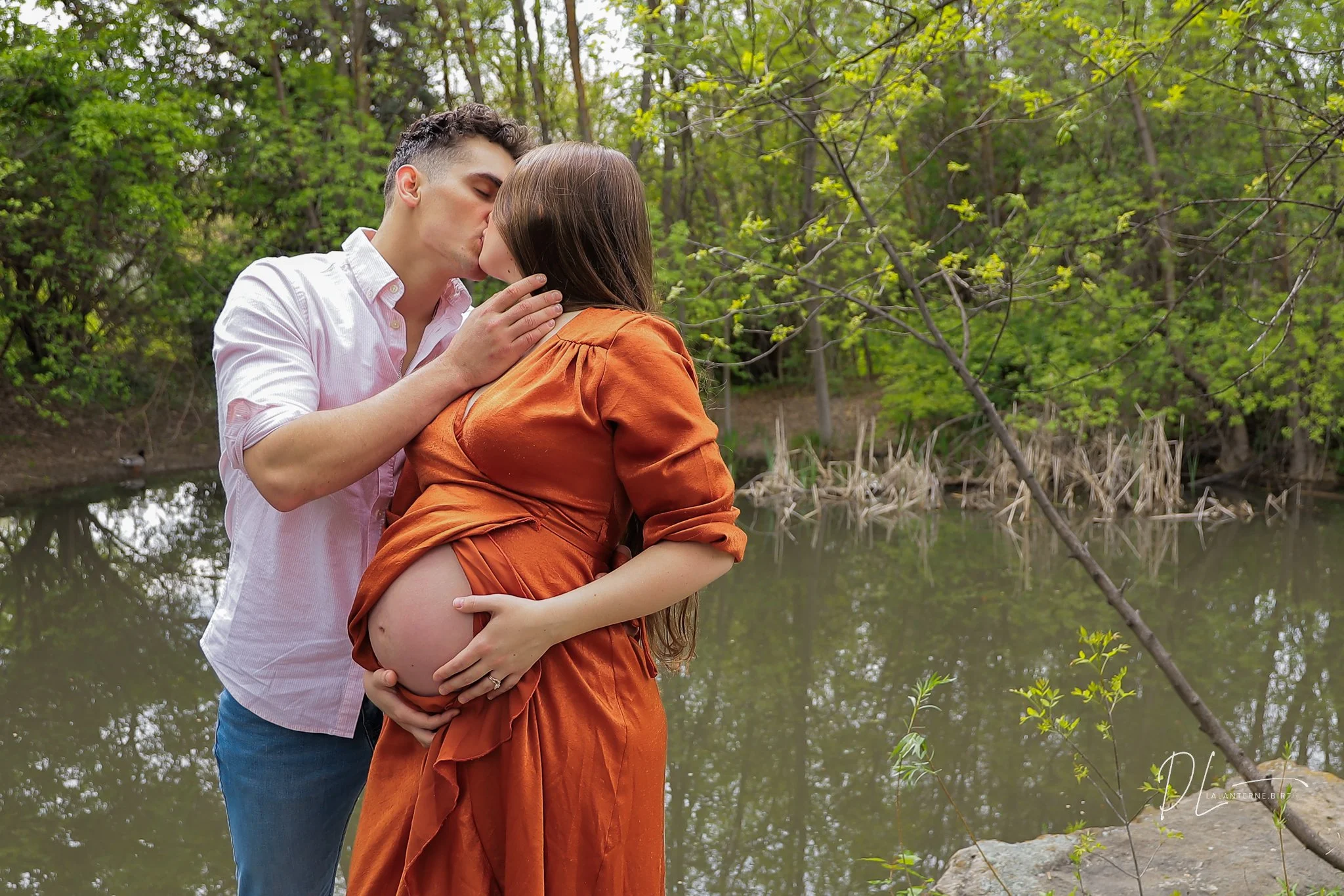A pregnant woman in an orange dress and a man are sharing a kiss outdoors by a pond, with trees and greenery in the background.