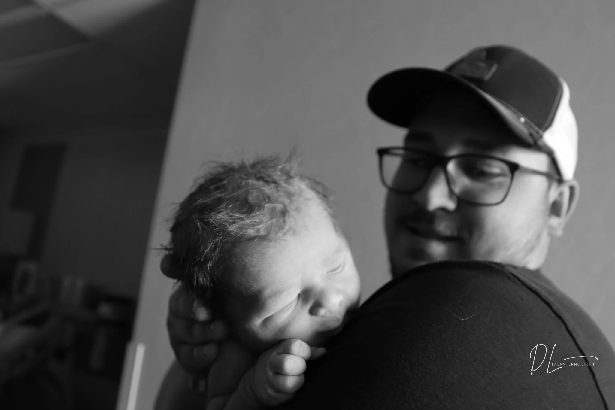 A man wearing glasses and a baseball cap holding a newborn baby close to his shoulder in a black and white photo.