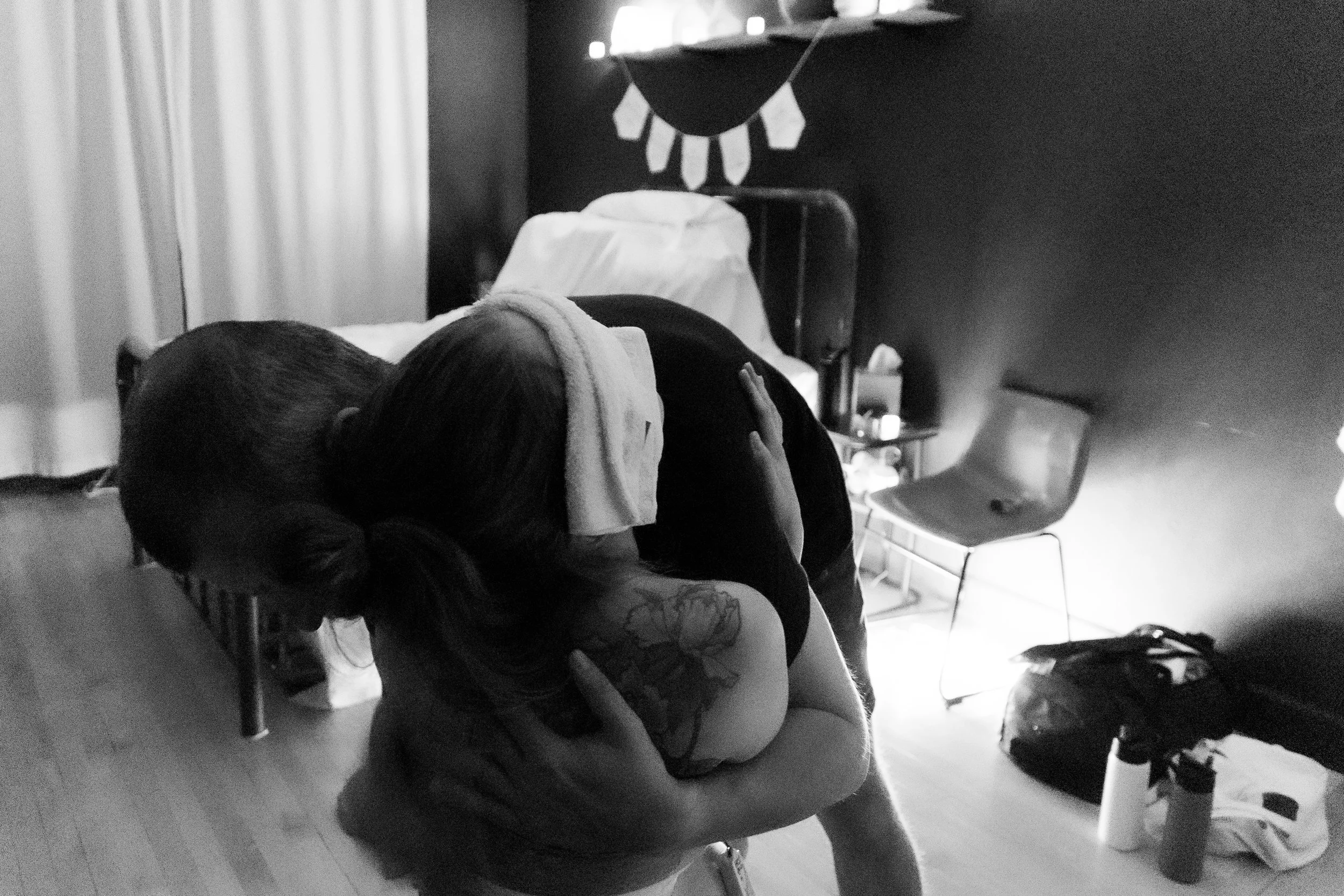 Two people hugging each other in a room, one with a towel on their head, in front of a bed in a dimly lit room.